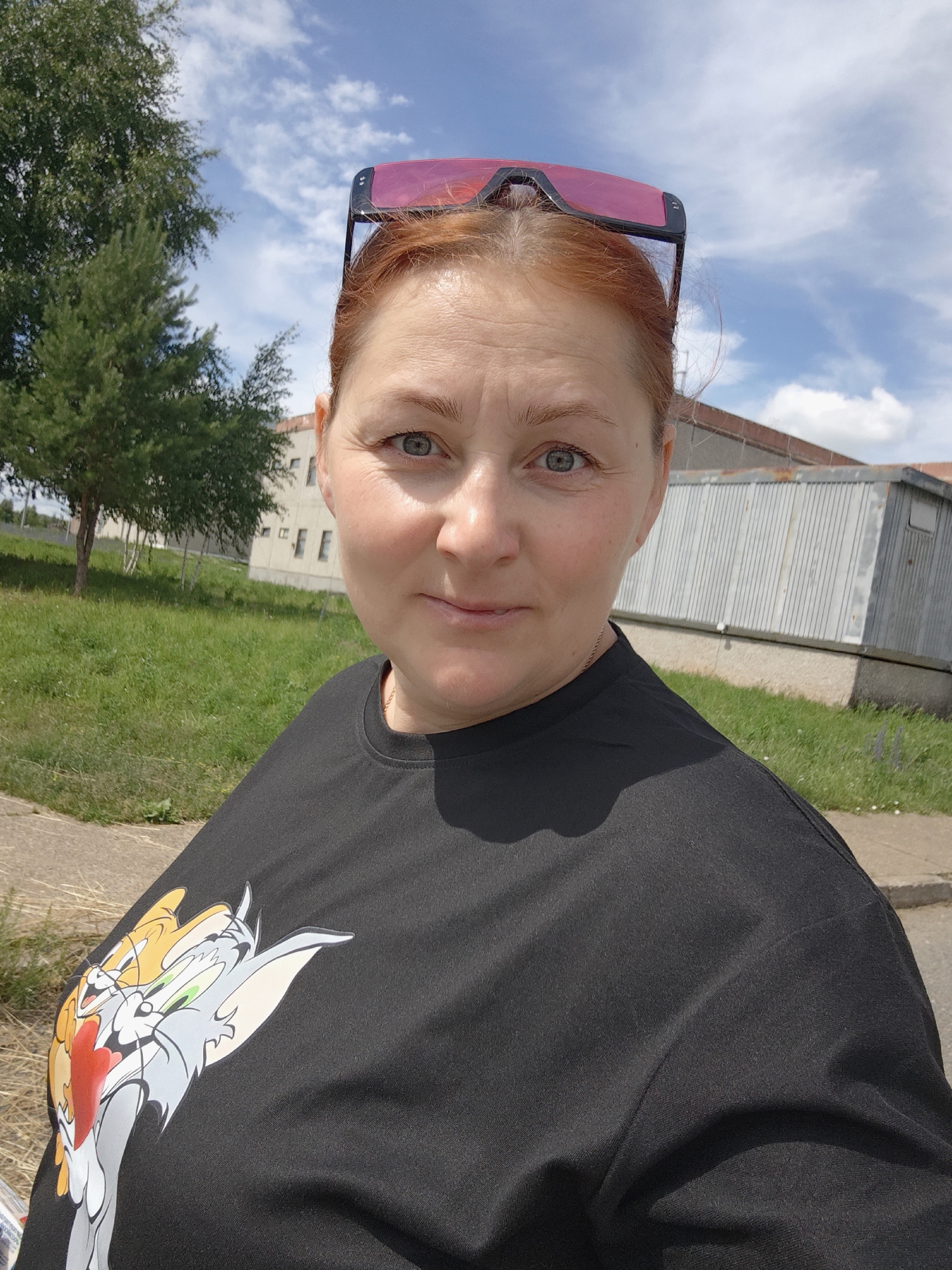 A woman with red hair pulled back and pink sunglasses resting on her head is shown in a close-up selfie taken outdoors. She is wearing a black t-shirt with a cartoon image of Tom and Jerry on the left side. The background includes a green grassy area, a tree, and a light-colored building with windows. The sky is blue with scattered clouds. The scene is likely located in the city of Марковский, Russia, based on the provided context.