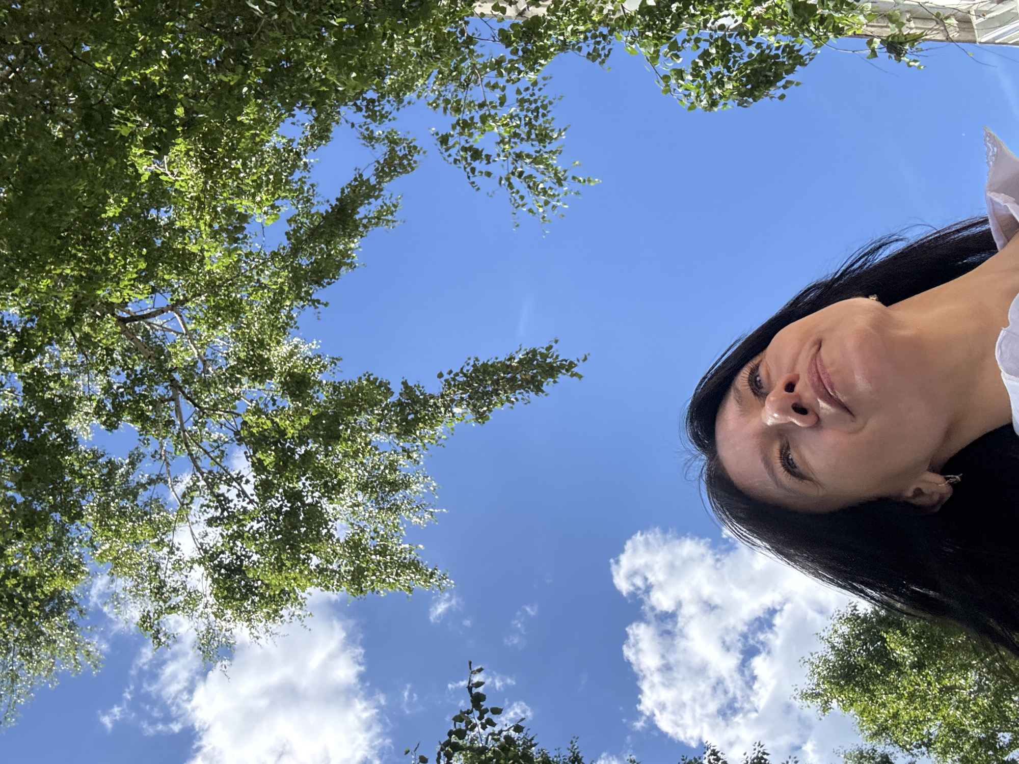 A low-angle, full-frame shot shows a woman with long, dark hair looking up at the sky, framed by the leaves of birch trees. The sky is a vibrant blue with scattered white clouds. The woman's face is partially illuminated by the sun, highlighting her features. The scene appears to be set outdoors in a location with ample greenery, possibly a park or a tree-lined street in Chaikovsky, Russia. The overall impression is one of serenity and connection with nature.