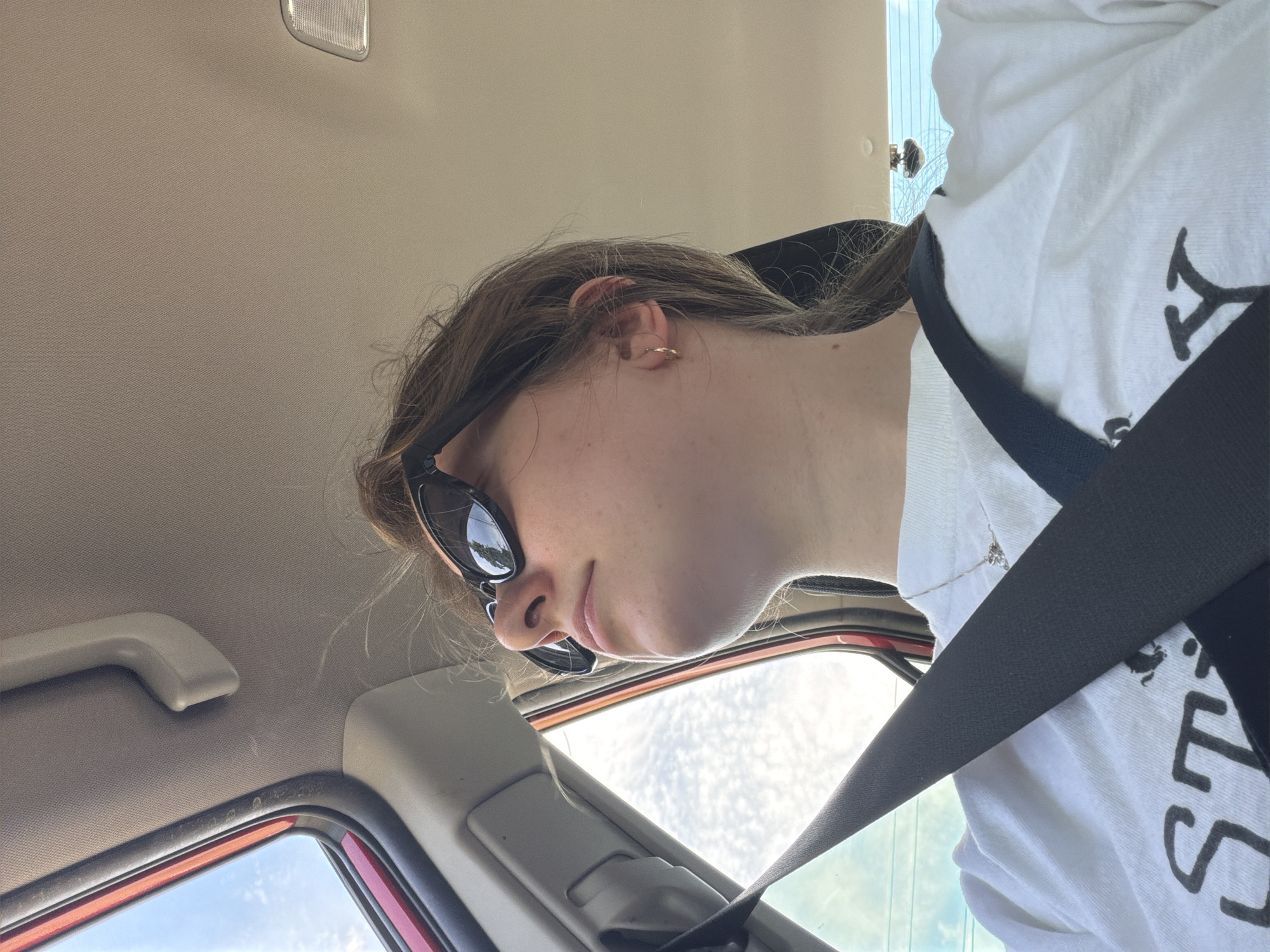 A young person with brown hair, wearing sunglasses and a white t-shirt with a graphic design, is looking up and to the right inside a vehicle. They are wearing a seatbelt. The interior of the car is visible, including the ceiling and a portion of the window. The location is Beaverton, United States of America.