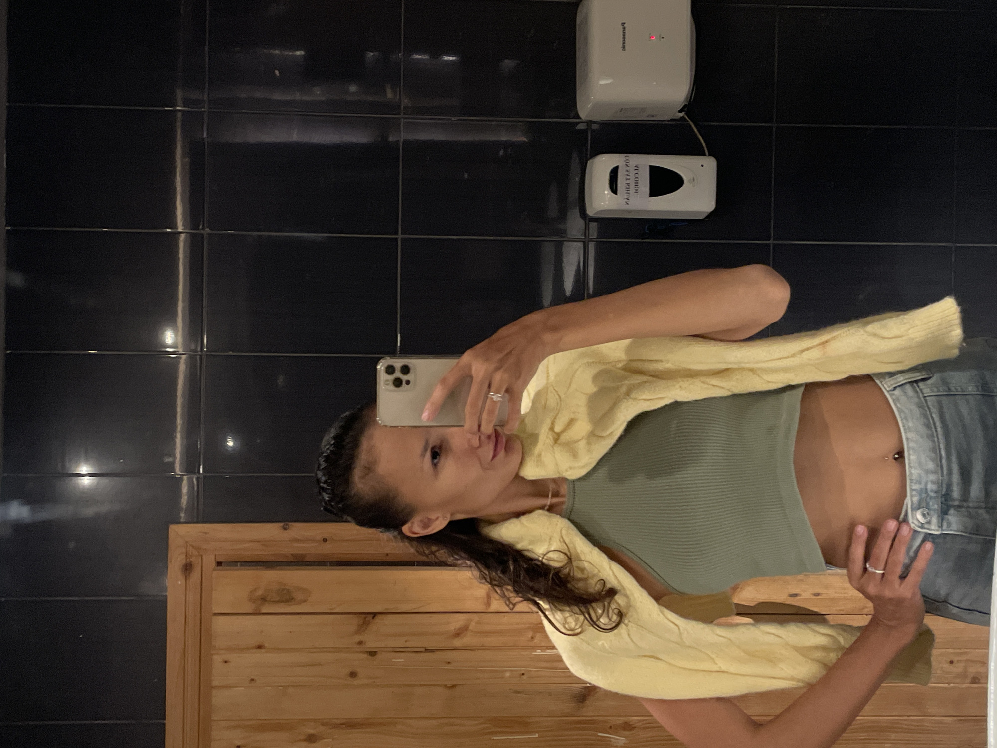 A young woman is taking a mirror selfie in an indoor setting. She is wearing a light green ribbed crop top, light blue denim jeans, and a light yellow knitted cardigan draped over her shoulders. She has dark hair styled back, and rings are visible on her fingers, along with a navel piercing. Her smartphone, an iPhone with multiple cameras, is held up to capture the reflection.
The background features a wall composed of dark, glossy black tiles. In the upper right quadrant of the image (when oriented correctly), two white wall-mounted dispensers are visible. The top dispenser has "Panasonic" text and a small red light, likely a hand dryer or soap dispenser. Below it, a second dispenser, reflecting reversed text, displays "ALCOHOL" and "HAND SANITIZER," indicating it is a sanitizer dispenser. The lower portion of the scene is a light brown wooden slatted partition or wall. The setting appears to be a public or commercial restroom or changing area, consistent with the location in Phu Thuy Ward, Vietnam.