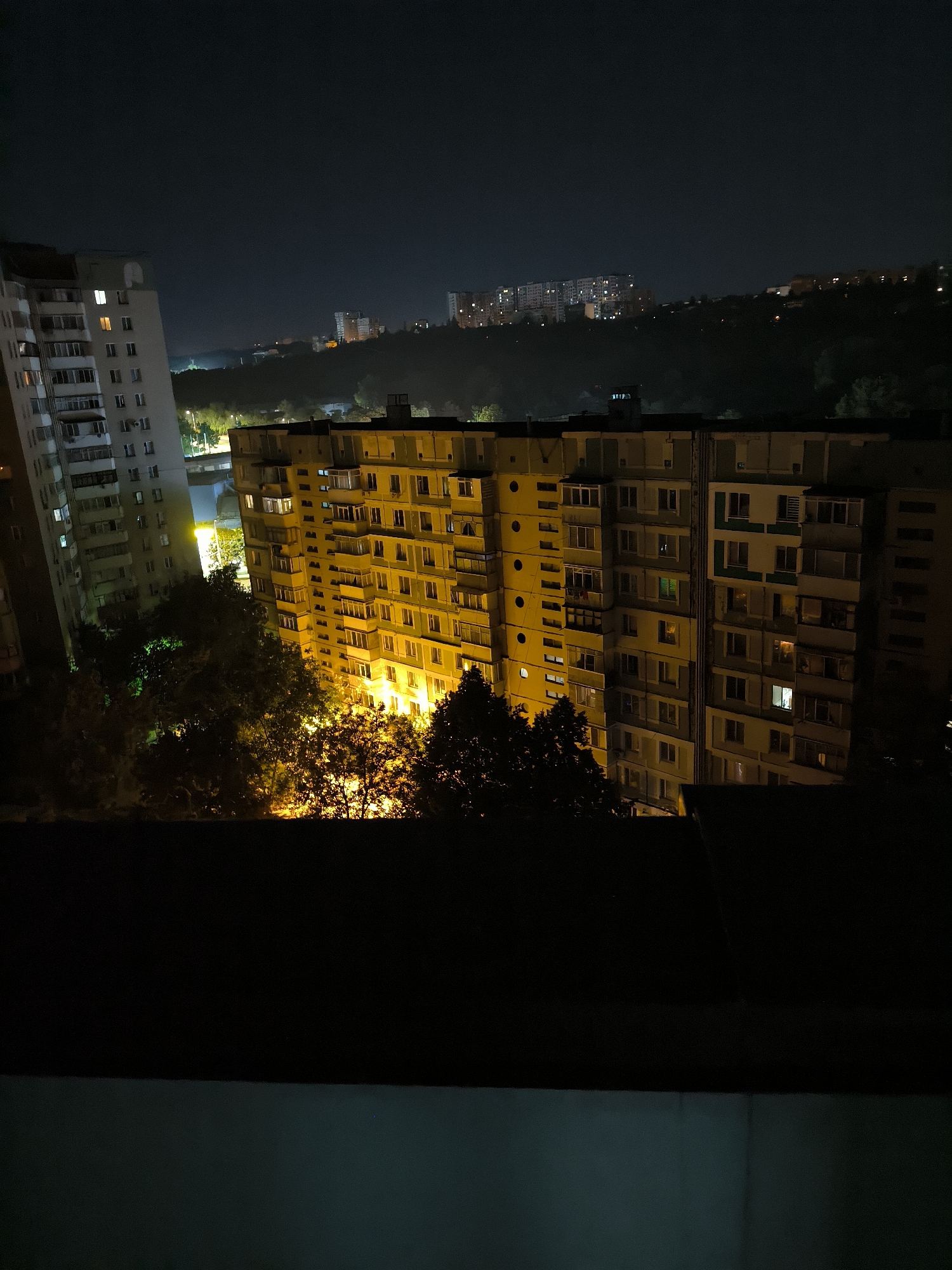 The image captures a nighttime view of a residential area in Chișinău, Moldova, likely from a higher vantage point, possibly a balcony or an upper floor. In the foreground, the dark silhouette of a structure, possibly a building's roof or balcony railing, obscures the bottom portion of the frame.
Dominating the mid-ground are two large, multi-story apartment buildings. The building on the left is lighter in color, with visible windows illuminated from within by a warm, yellowish light. This building appears to be slightly angled, creating a dynamic visual. To its right is a larger, yellowish apartment block. Its numerous windows are also lit, with some glowing more intensely than others, suggesting varying levels of occupancy or activity within the apartments. The architecture of this building features repetitive balconies and window designs, typical of Soviet-era apartment blocks.
In front of and between these buildings, several trees are visible. Their dark foliage is partially illuminated by the artificial lights from the apartments, creating pockets of orange and green glow amidst the shadows.
Further in the background, the hilly terrain of Chișinău is apparent. More apartment buildings and structures are scattered across the distant hills, their windows appearing as scattered points of light against the dark night sky. The overall scene conveys a sense of urban life continuing after dark, with the lights from the residential buildings piercing the night. The atmosphere is quiet and still, characteristic of a residential neighborhood at night.