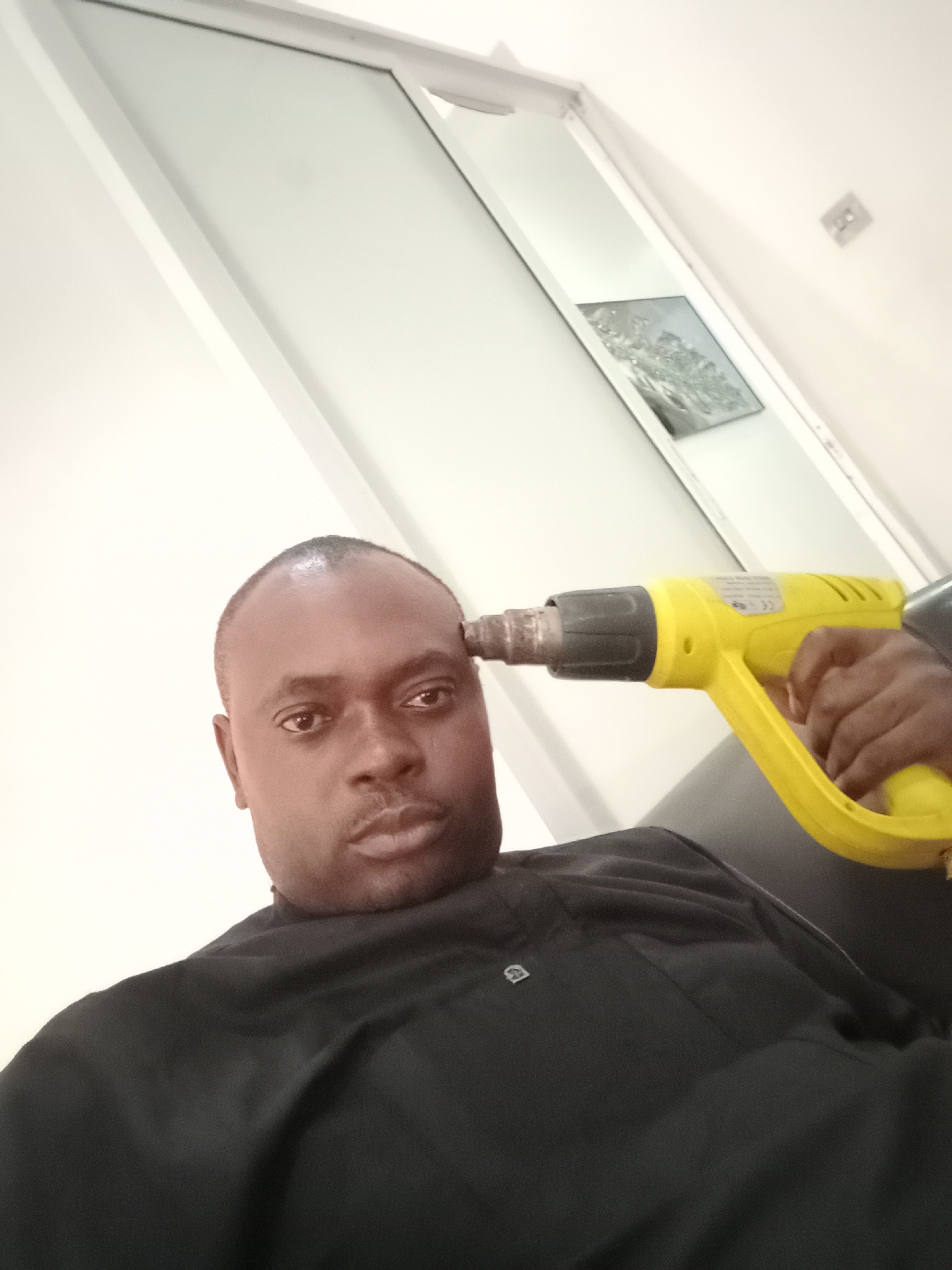 A man is holding a yellow heat gun to his forehead. He is wearing a black shirt. The background consists of a white wall, a door, and a framed picture. There is no evidence to suggest that this is in Gwarinpa, Nigeria.