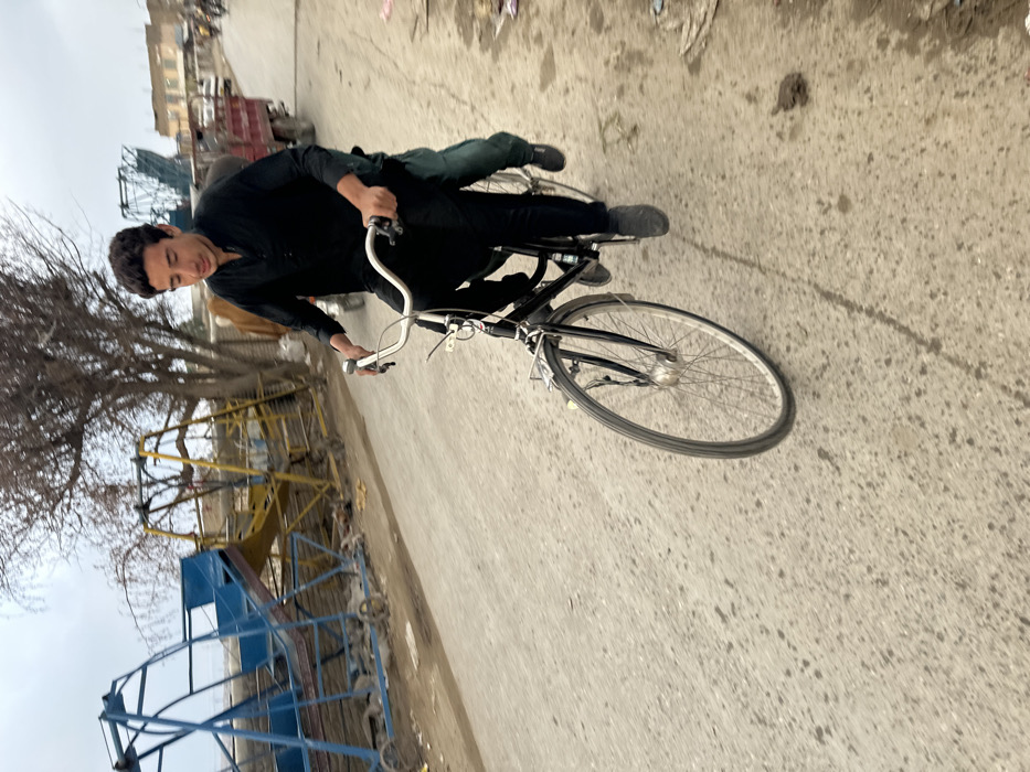 This image captures a street scene in Quetta City Tehsil, Pakistan, depicting a young male actively riding a black road bicycle. The cyclist, dressed in a black shalwar kameez and dark shoes, holds the handlebars while looking slightly downward. A second individual, partially obscured behind the cyclist, is visible by their left leg clad in green/blue trousers and a dark shoe, indicating they are also on the bicycle. The bicycle features silver handlebars, a front fender, and a small luggage rack. The street surface is paved and exhibits wear, with visible dirt and minor imperfections. In the left background, two large, disused or stored open metal structures, one blue and one yellow, are visible next to a bare, deciduous tree. Further in the background on the right, buildings and a red truck are discernible. The sky is overcast, providing diffused lighting.