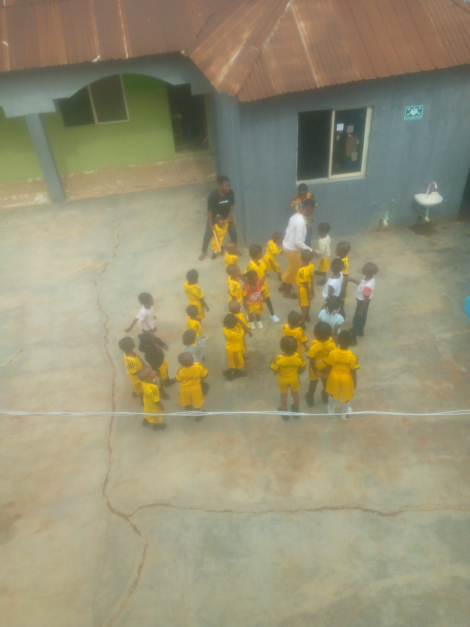 The image depicts a group of young children gathered in an outdoor area, possibly a schoolyard or playground, in an urban location identified as Unknown, Unknown. The children are mostly wearing yellow uniforms, and some are also dressed in white or red. Adults are present, presumably teachers or caretakers. The setting includes buildings with distinct architectural features, such as rusted corrugated roofs and variations in paint colors. A washbasin is visible near the building. The environment is characterized by a concrete surface with noticeable cracks. The aerial view suggests the photo was taken from an elevated position.