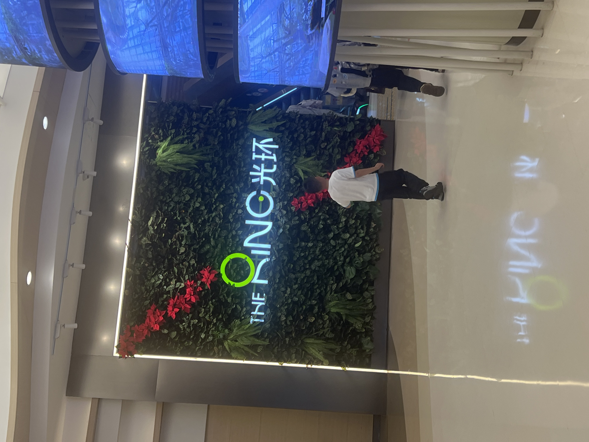A vertical shot shows a green wall at the entrance to “The Ring” shopping mall in the Liangjiang district of Chongqing, China. The wall is covered with green plants, with patches of bright red poinsettias. The mall’s logo, “The Ring 光环” is vertically spelled out in neon letters, the “O” in the logo shaped as a ring of light. A young boy wearing a white shirt and dark pants stands facing the wall, slightly obscuring the logo. The floor of the mall is pale and reflective, with a mirrored image of the logo. Above the wall are large, circular screens displaying blue light. In the background, a group of people can be seen walking past, their figures blurred. The environment is brightly lit and appears to be indoors.