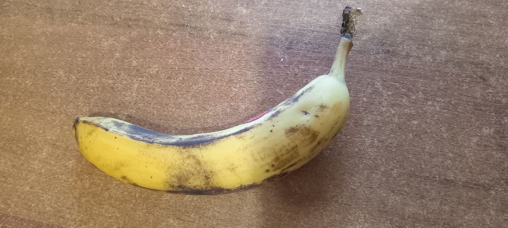 A single, ripe yellow banana with brown spots is lying on a textured, brown surface. The banana is curved and lies on its side, with the stem pointing to the lower right. The surface appears to be a wooden table or countertop. The lighting is consistent and illuminates the banana and the surface evenly. There are no people or other objects visible in the image.