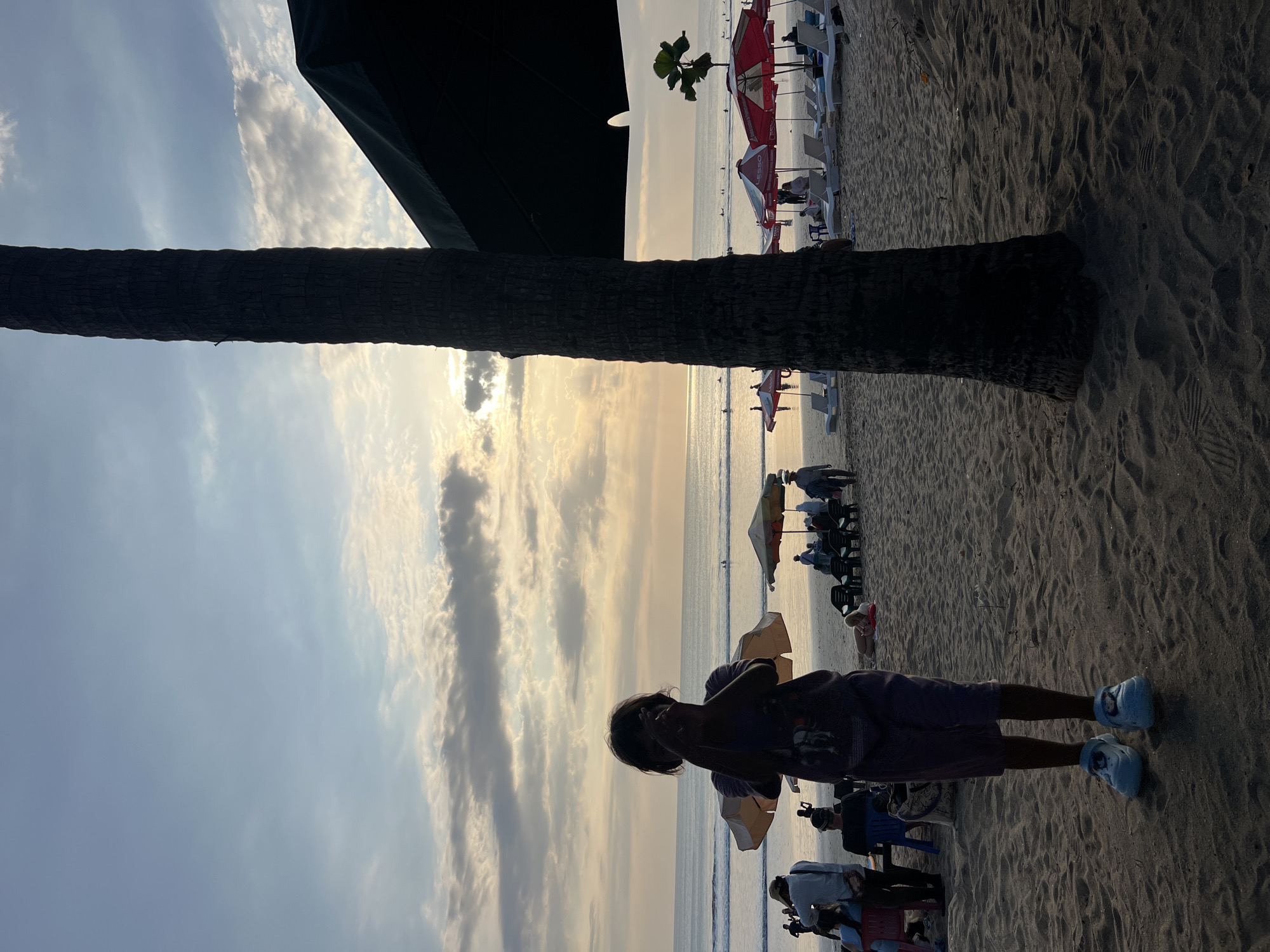 A young child stands on the sandy shore of Kuta Beach, Indonesia, holding two open umbrellas behind their shoulders. The child is wearing a dark t-shirt and shorts, with light-colored shoes. The beach is populated with other people, some sitting on lounge chairs under umbrellas, while others are walking or standing. The ocean is visible in the background with small waves and several surfers. The sky is partly cloudy with the sun setting, casting a warm glow over the scene. A large palm tree trunk is prominently positioned in the foreground, partially obscuring the view of the beach. Red and white umbrellas are set up along the shoreline, and lounge chairs are arranged on the sand. The overall atmosphere is that of a relaxed, late afternoon at a popular tourist destination.