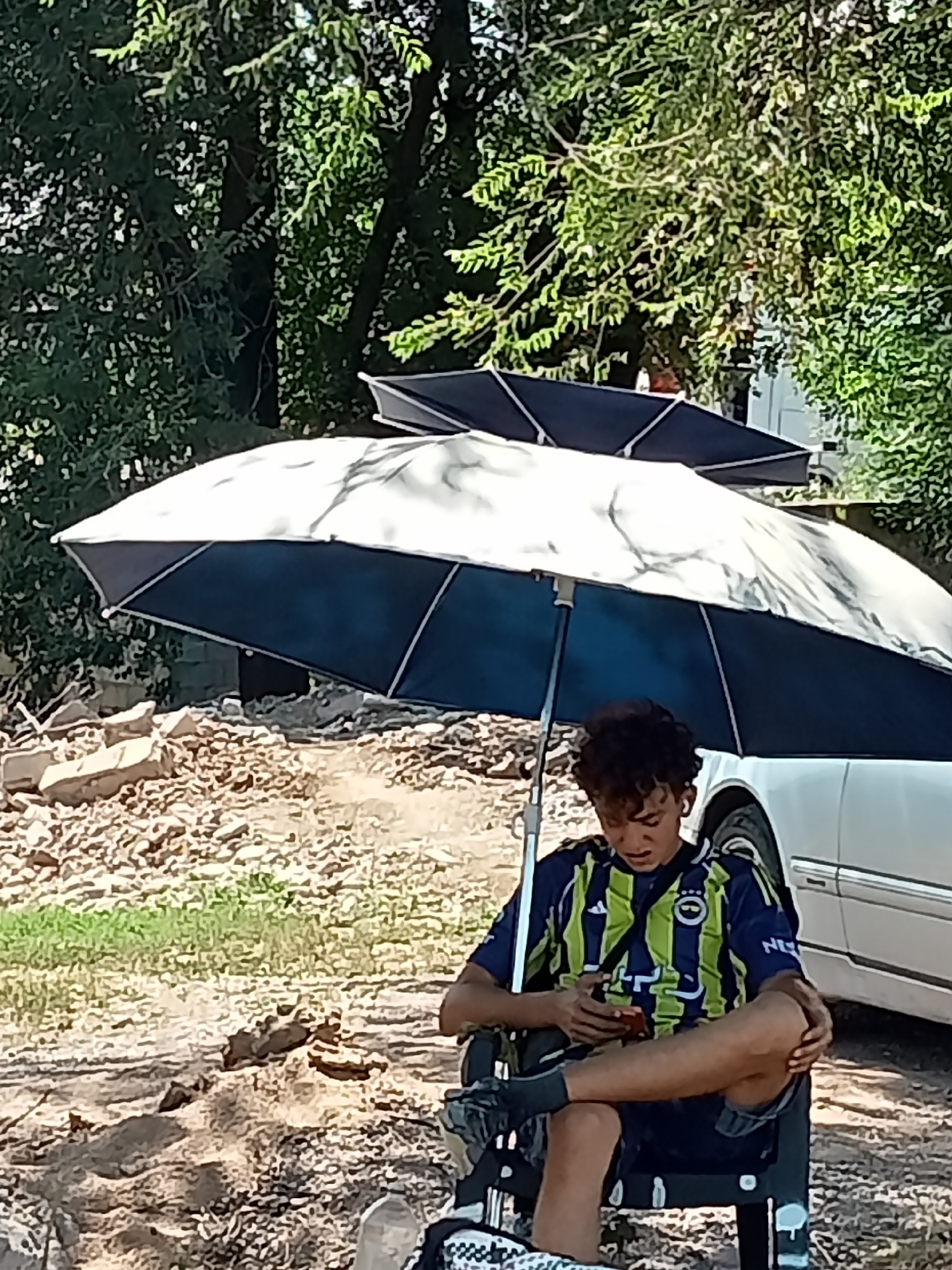 In Qaskelen, Kazakhstan, a young person with curly hair sits in a folding chair under a large umbrella. The individual is wearing a navy blue and lime green striped athletic shirt and is looking down at a mobile phone in their hands. Their legs are crossed, and one foot is resting on the chair's leg. Behind the person, a white car is partially visible, along with a pile of debris that appears to be rocks and concrete. The environment is outdoors, with trees and foliage in the background, suggesting a park or a somewhat undeveloped area. Another umbrella, dark blue and partially open, is visible behind the main umbrella. The ground is covered in dirt and small stones.