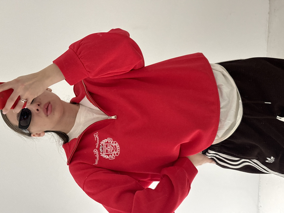 A single individual, appearing to be female, is depicted taking a mirror selfie indoors against a plain white wall. The specific location within the city of Krasnodar, Russia, cannot be determined from the visual evidence.
The individual is wearing a red quarter-zip sweatshirt featuring white embroidery on the left chest, which includes a crest-like emblem with the text "CREATION GHD CREATED 0210 SMART PLAY MMXXII" and "Dream unlimited" above it. A white undershirt is visible beneath the red sweatshirt. The lower body is dressed in black Adidas track pants with white stripes running down the sides and a white Adidas logo visible on the left leg.
The individual wears black, rectangular-framed sunglasses. A silver-colored ring is present on the fourth digit of the left hand. A red smartphone, consistent with an Apple iPhone, is held in the left hand and partially obscures the individual's face, indicating the act of capturing a reflection (selfie). The individual's right hand is placed on their right hip. Their lips are slightly parted. No other individuals are present, and there are no discernible interactions with external subjects or objects beyond the smartphone. The environment is uniformly white and devoid of distinguishing features.