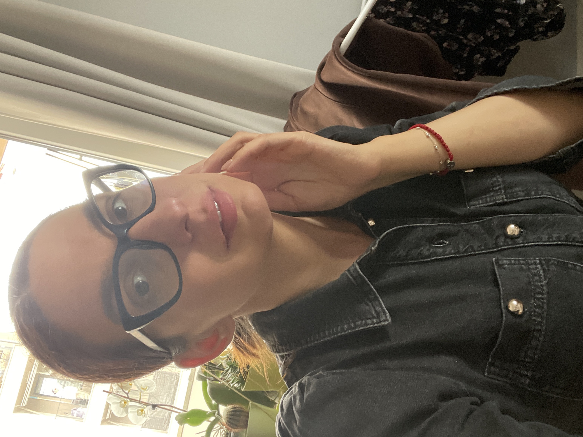 A selfie photograph shows a person wearing glasses and a black denim jacket. The person has a red beaded bracelet on their left wrist. The background includes a window with white blinds, a clothes hanger with clothing, and green plants. The location is difficult to determine based on the provided visual information and, therefore, cannot be identified specifically as Sofia, Bulgaria.