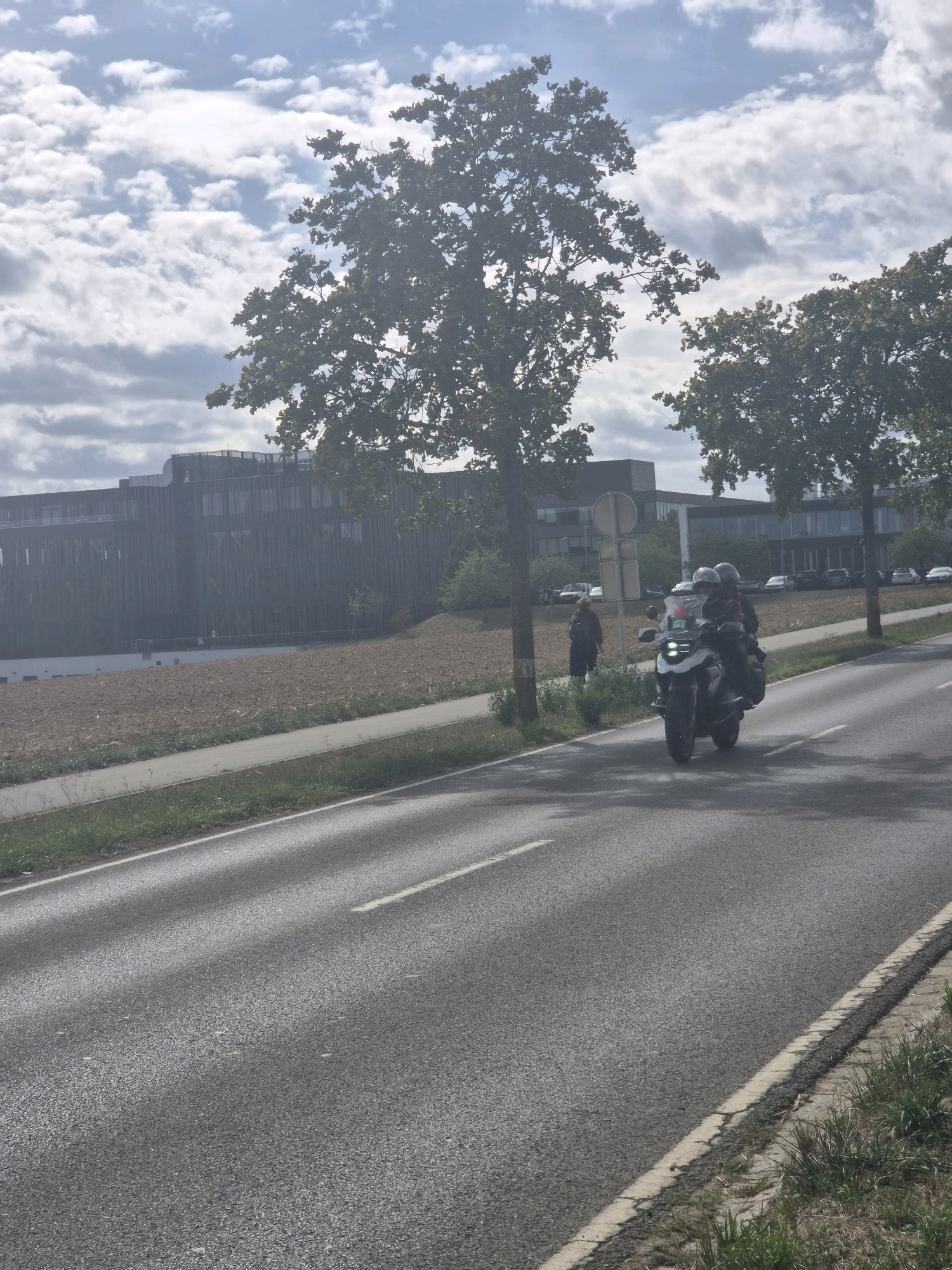 The image shows a motorcycle with two riders on a road in Leudelange, Luxembourg. The motorcycle is white and black, and the riders are wearing helmets and dark clothing. The road is asphalt, with white lane markings. On the left side of the road, there is a sidewalk and a grassy area. In the background, there is a modern office building with a dark facade and a large field. The sky is partly cloudy, with white clouds against a blue sky. The sun appears to be shining, casting shadows on the road. There are also trees visible on the left side of the road and in the background. The image appears to have been taken in the daytime.