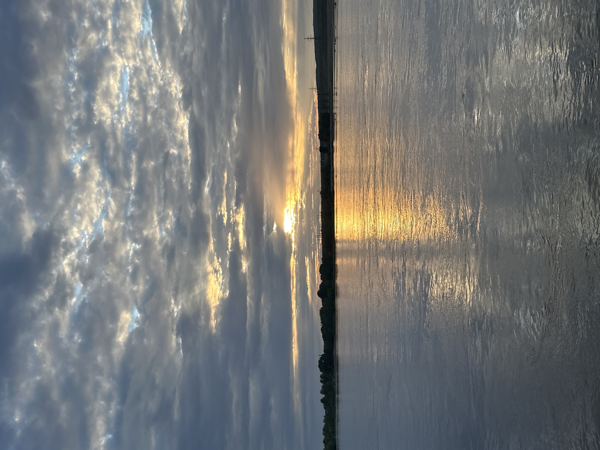 The image shows a body of water with a reflection of the sunset on its surface. The sky is cloudy with the sun breaking through the clouds, casting rays of light. A treeline with some structures can be seen in the distance across the water. Several small boats are visible on the water near the treeline. Power line towers are also present on the horizon.