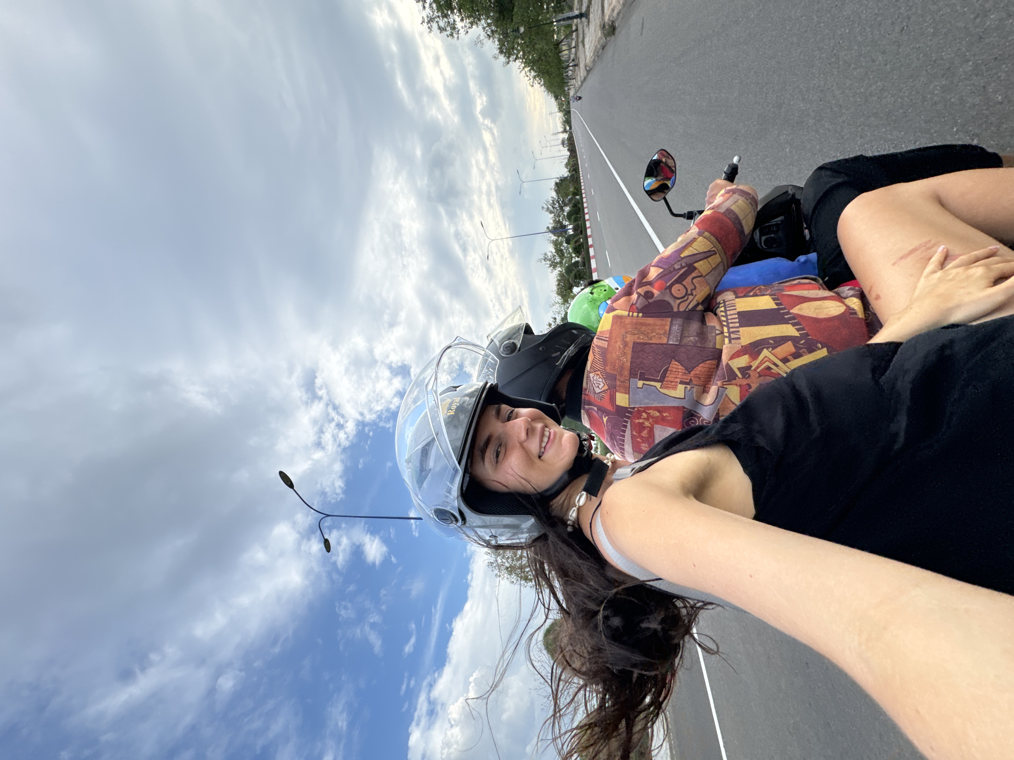 This image captures a selfie taken by a young woman riding as a passenger on a scooter or motorcycle on an open road. The woman, positioned in the lower-left foreground, smiles brightly at the camera. She wears a grey helmet with a clear visor, on which the brand "Royal" is visible, and has long, dark hair flowing freely. Her attire includes a black tank top under a black short-sleeved top, and a beaded necklace. Her left hand rests on her thigh, which shows some faint scratches or marks.
The driver of the scooter is partially visible to her side, wearing a colorful, intricately patterned long-sleeved jacket and a green helmet. The setting is an asphalt road, marked with white lines and red-and-white curb markings, stretching into the distance. Tall streetlights with curved arms line the side of the road, alongside green trees and other vegetation. The sky above is prominent, featuring a mix of bright blue patches and large, fluffy white and grey clouds, indicating a daytime scene with partly cloudy weather. Another distant vehicle can be faintly observed down the road. The image conveys a cheerful moment of travel, likely in Phu Thuy Ward, Vietnam.