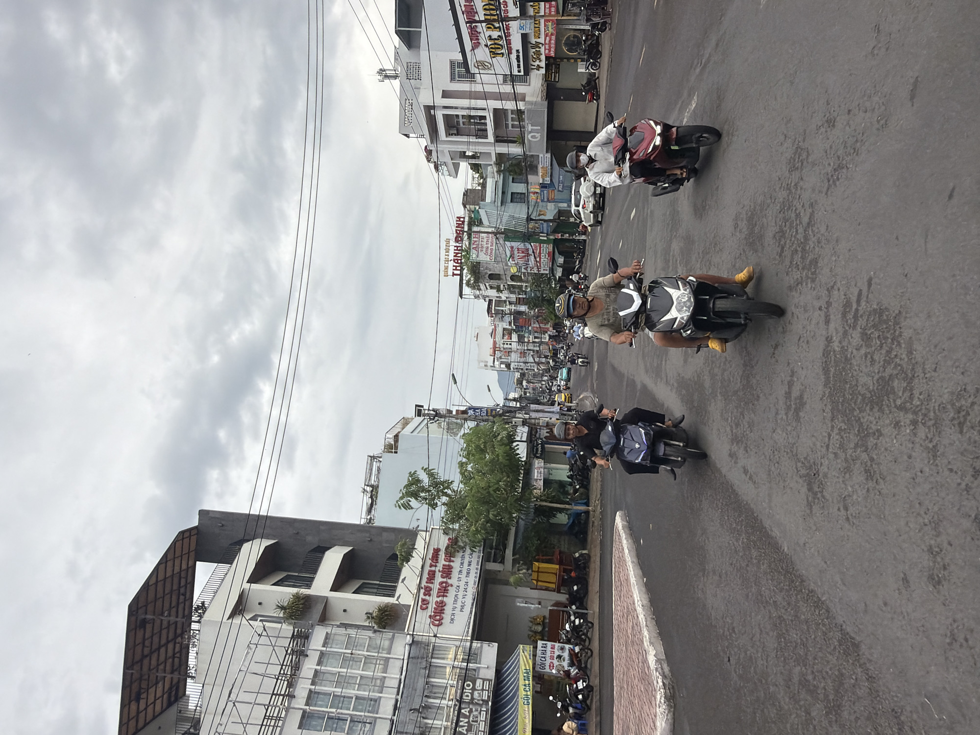 The image depicts a street scene in Nha Trang, Vietnam, characterized by a cloudy sky and buildings lining both sides of a paved road. Three individuals are riding motorcycles in the foreground, moving from left to right.
The leftmost motorcyclist is a man wearing a dark shirt and pants, riding a dark-colored scooter. He is looking forward, focused on the road.
In the center, another man is riding a black scooter, wearing a light-colored t-shirt with a pattern and dark shorts. He is wearing a helmet and yellow shoes, and his gaze is also directed forward.
To the right, a third individual, also on a red scooter, is wearing a white long-sleeved shirt and dark pants. This person is wearing a face mask and a helmet.
The buildings on the left side of the street are a mix of modern and older architectural styles, some with balconies and visible windows. Signs in Vietnamese are visible on some of these structures, including one that reads "CÔNG THỌ SẨU PHONG" and another with "AN AUDIO". Greenery is present, with trees and potted plants visible.
On the right side of the street, more buildings are visible, also featuring various architectural designs. Signs in Vietnamese, such as "THÀNH DANH" and "TOC PHONG", are present. Electrical wires are strung overhead, a common feature in urban Vietnamese streetscapes. The road itself is asphalt, with painted lane markings. In the background, the street extends into the distance, with more buildings and vehicles visible. The overall atmosphere suggests a typical day in a bustling Vietnamese city.