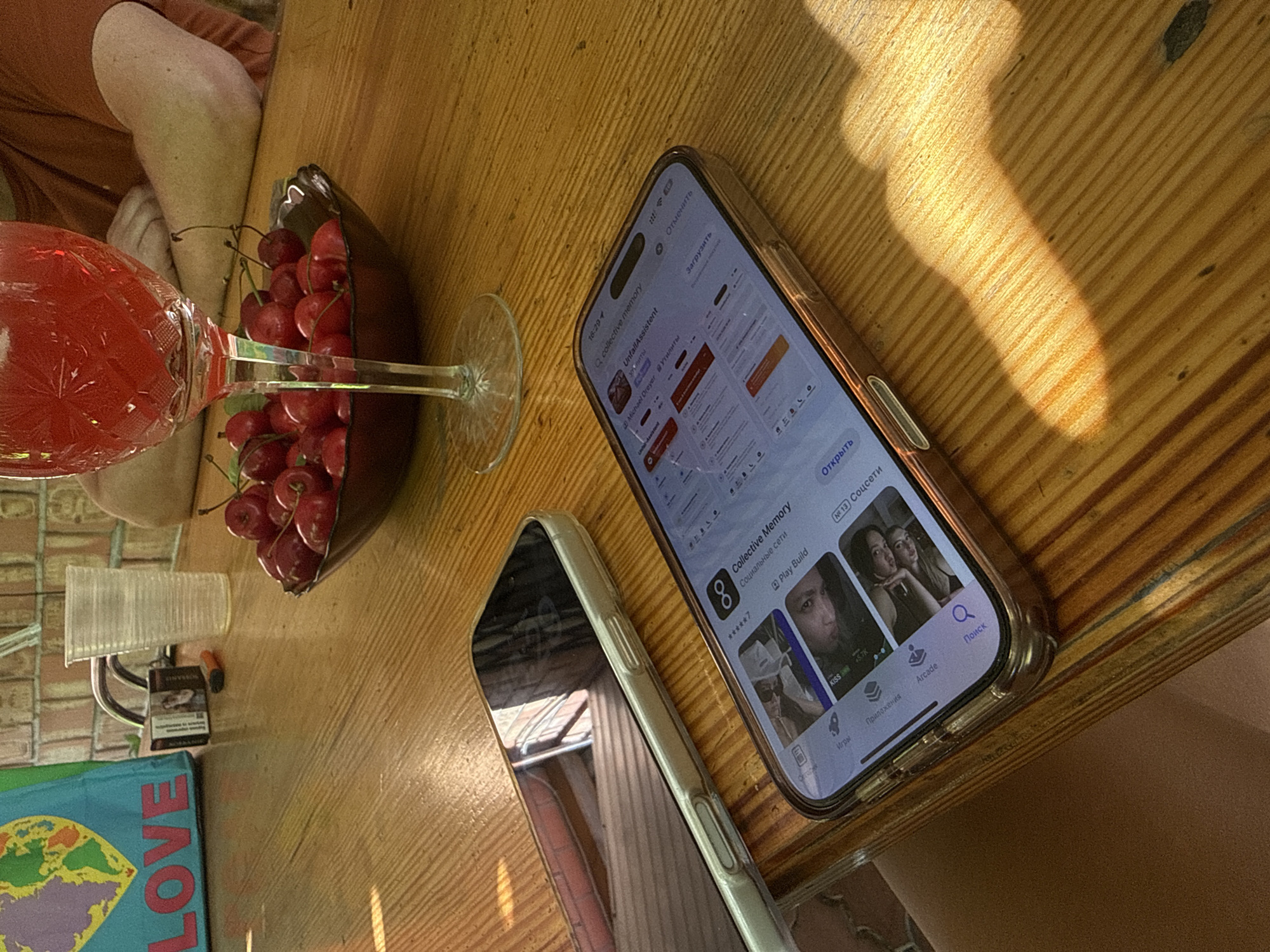 This is a medium-angle, eye-level shot of two smartphones lying on a wooden table. In the background, a glass of red liquid, a bowl of cherries, a plastic cup, and a pack of cigarettes are visible. The overall scene appears to be outdoors, likely in a setting like Obukhivka, Ukraine, given the presence of the "LOVE" bag and the brick wall, suggesting a domestic or recreational environment. The smartphones are the primary focus, with one displaying an app interface related to social media or memory storage, and the other's screen reflecting the surrounding environment.