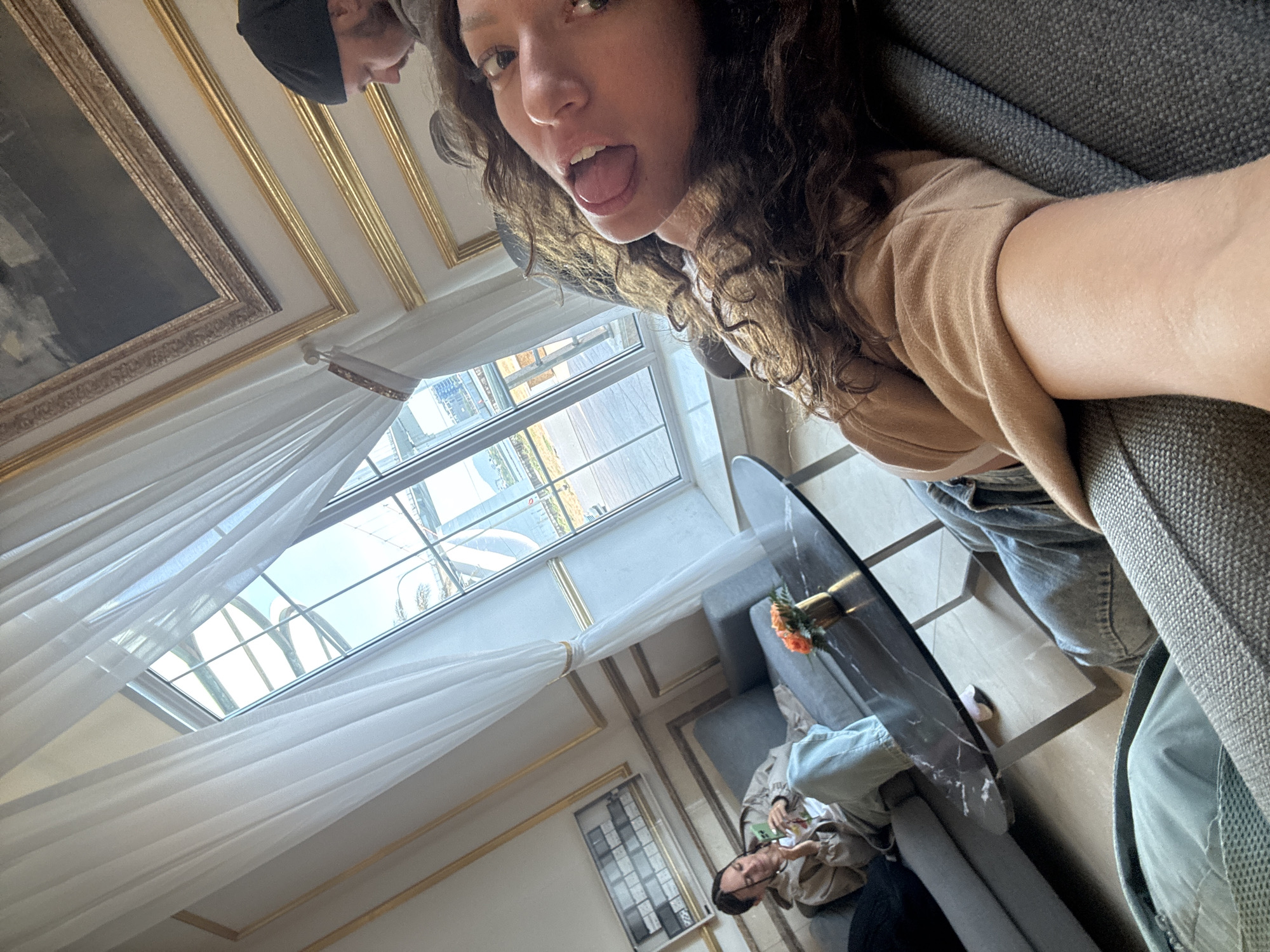 The image shows two people in a room with ornate molding on the walls and ceiling. One person is holding the camera at an angle, showing their face and part of their body. They have long, curly hair, a tan shirt, and jeans. Their mouth is open, and they are looking at the camera. Another person is sitting on a gray sofa and looking at something in their hands. A black coffee table with a vase of flowers is in front of them. A large window with white curtains is visible in the background, showing a view of a cityscape with buildings and a body of water. A large painting hangs on the wall. The location is Đà Nẵng, Vietnam, based on the architectural style.