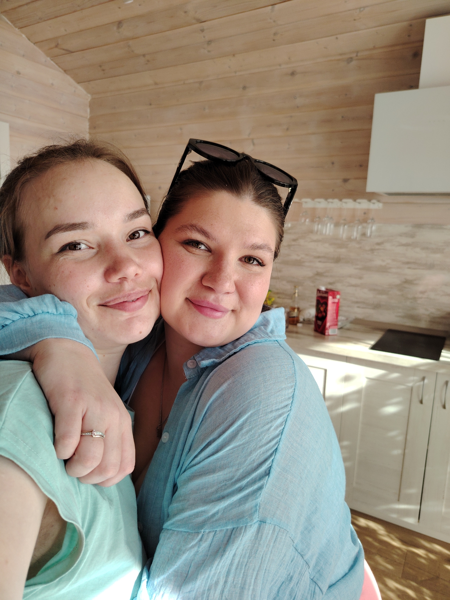 Two women are embracing indoors. The woman on the left has light brown hair pulled back from her face, fair skin, and is wearing a light green shirt. She has a ring on her left ring finger. Her right arm is around the other woman's shoulders. The woman on the right has darker hair, pinned up with sunglasses perched on her head. She is wearing a light blue shirt. They are both smiling.
The background shows a kitchen setting. The walls are clad in light wood panels. In the background, there are white kitchen cabinets, a white range hood, and glasses hanging above the counter. On the counter, there is a small bottle and a carton of juice. The image appears to be taken in a residential setting. There is no evidence to suggest that this image was taken in Malyi Bukor, Russia, or any other specific location.