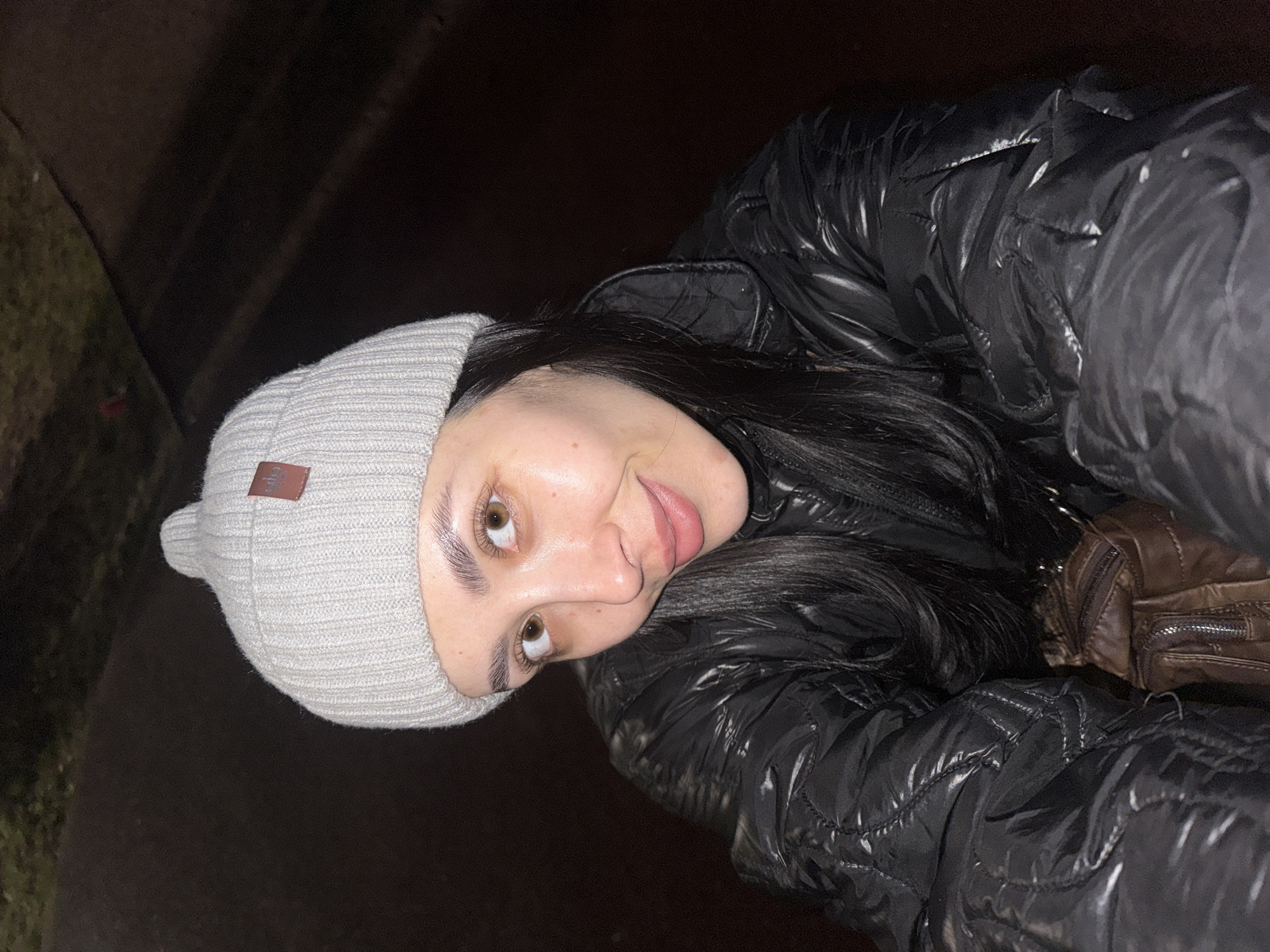A young woman is depicted in a close-up, sideways portrait, likely a selfie taken outdoors at night in Votkinsk, Russia. She is looking directly at the camera with a playful expression, her tongue slightly extended. Her eyes are light brown, and her eyebrows are dark and well-defined.
She wears a light grey ribbed knit beanie on her head, with a small, rectangular brown label visible on the side. The label features white text that appears to read "ALO". Her long, dark hair cascades down her back. She is dressed in a shiny, black puffer jacket, suggesting cool or cold weather. Part of a brown, leather-like bag or purse with a visible zipper is seen over her shoulder, just behind her jacket.
The background is very dark and indistinct, with faint hints of darker ground or foliage, indicating an outdoor setting with minimal light sources. The overall impression is a casual, nighttime portrait.