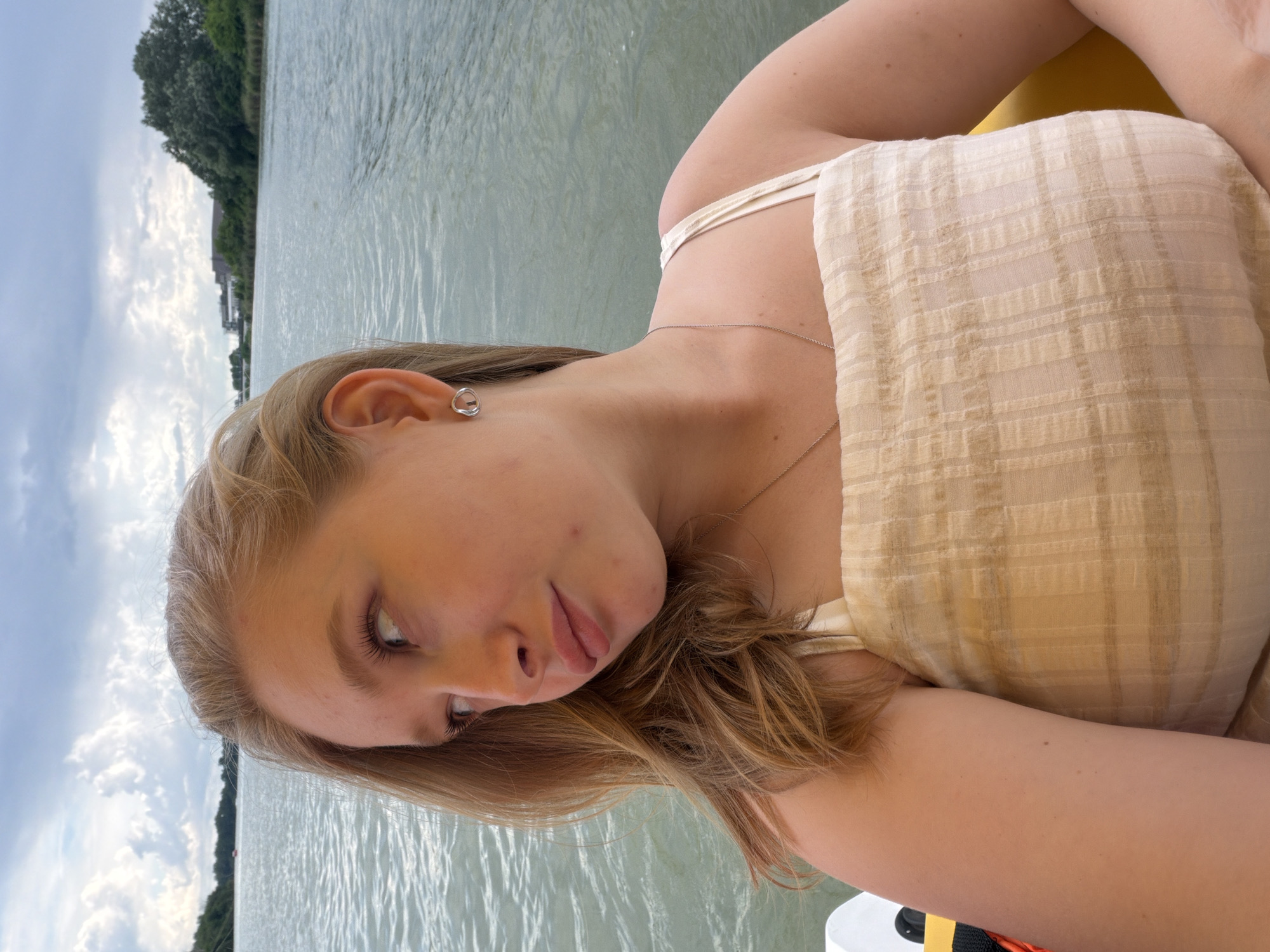 A young woman with blonde hair is photographed from the chest up, facing away from the camera and looking to her left. She is wearing a light-colored, textured top with thin straps and a silver necklace. She has on a small silver hoop earring in her visible ear. The background is a body of water with trees and buildings visible in the distance, under a cloudy sky. The location is not Krasnodar, Russia.