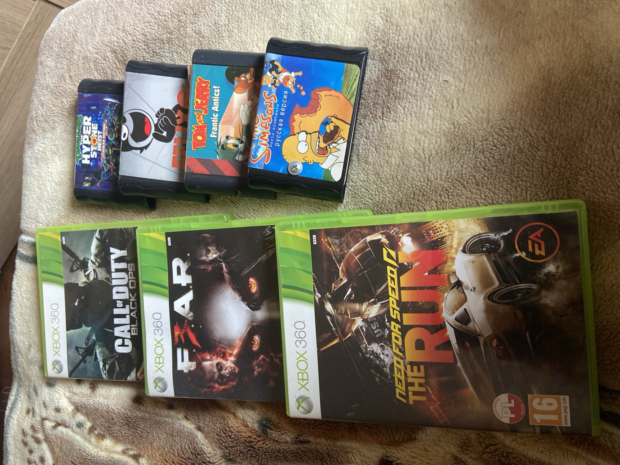 The image shows a collection of video game cartridges and Xbox 360 game cases lying on a brown and beige patterned surface, likely a bedspread or blanket. The cartridges include "Hyper Stone Heist," a black and white cartoon, "Tom and Jerry Frantic Antics!," and "The Simpsons" (in Russian). The Xbox 360 games are "Call of Duty: Black Ops," "F.E.A.R.," and "Need for Speed: The Run." The "Need for Speed" game has Polish age rating information.