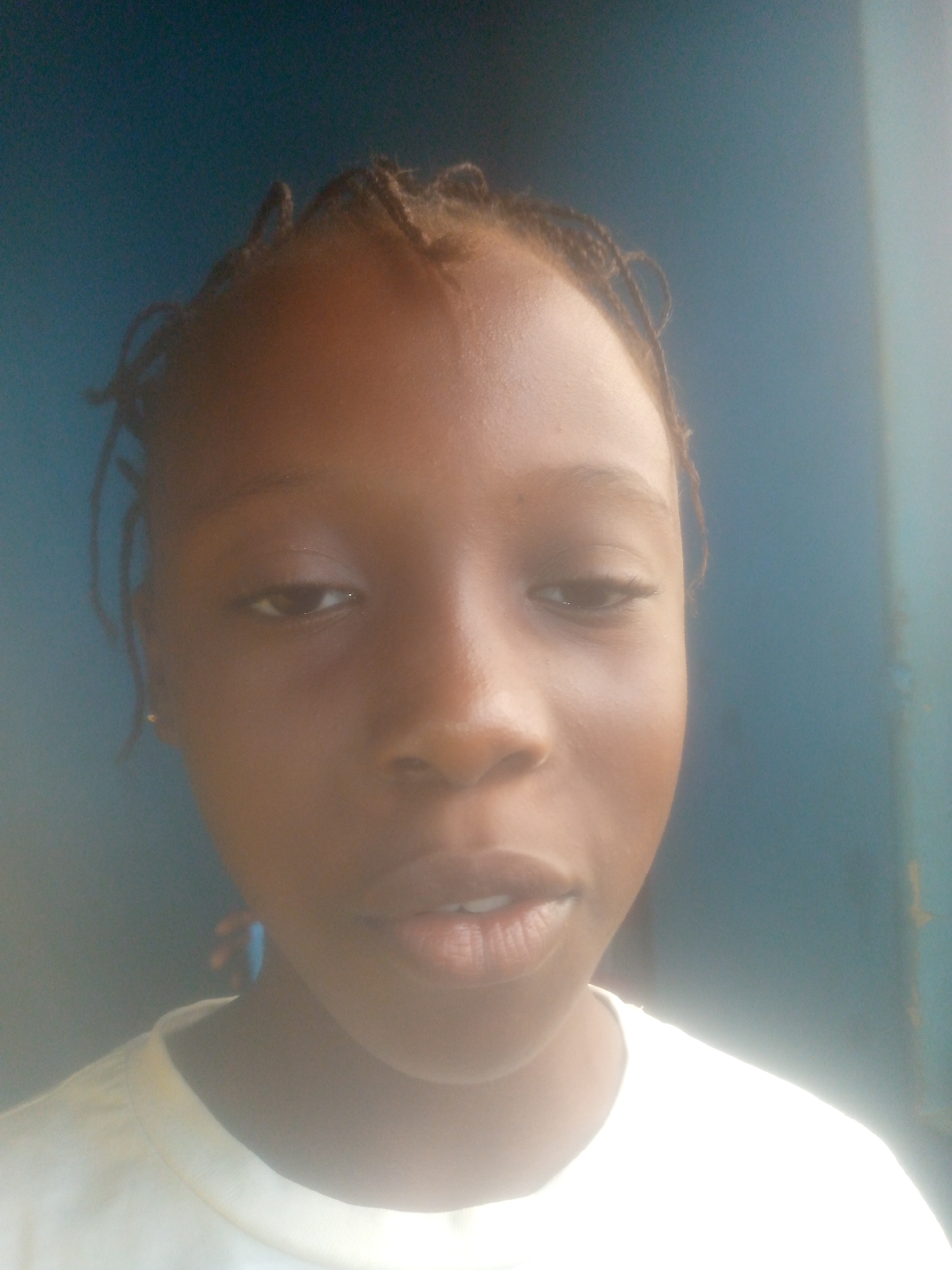 A close-up, slightly out-of-focus portrait of a young Black girl is presented against a blurred blue background. She has dark brown skin, a small nose, and lips that are slightly parted. Her hair is braided close to her scalp, with a few loose strands around her forehead. She is wearing a white t-shirt. The location is specified as the city of Unknown, Unknown.