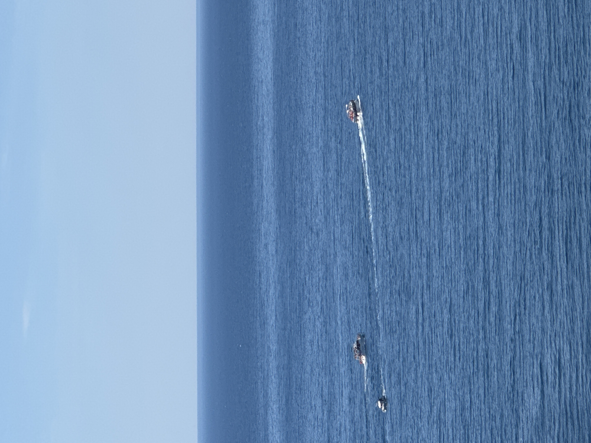 The image depicts a body of water, presumably the Adriatic Sea, near Sveti Stefan, Montenegro. The sky is visible on the left, with a gradient from light blue to a darker blue towards the center. Two boats are present in the sea. One is larger and further away. Both boats are creating wakes.