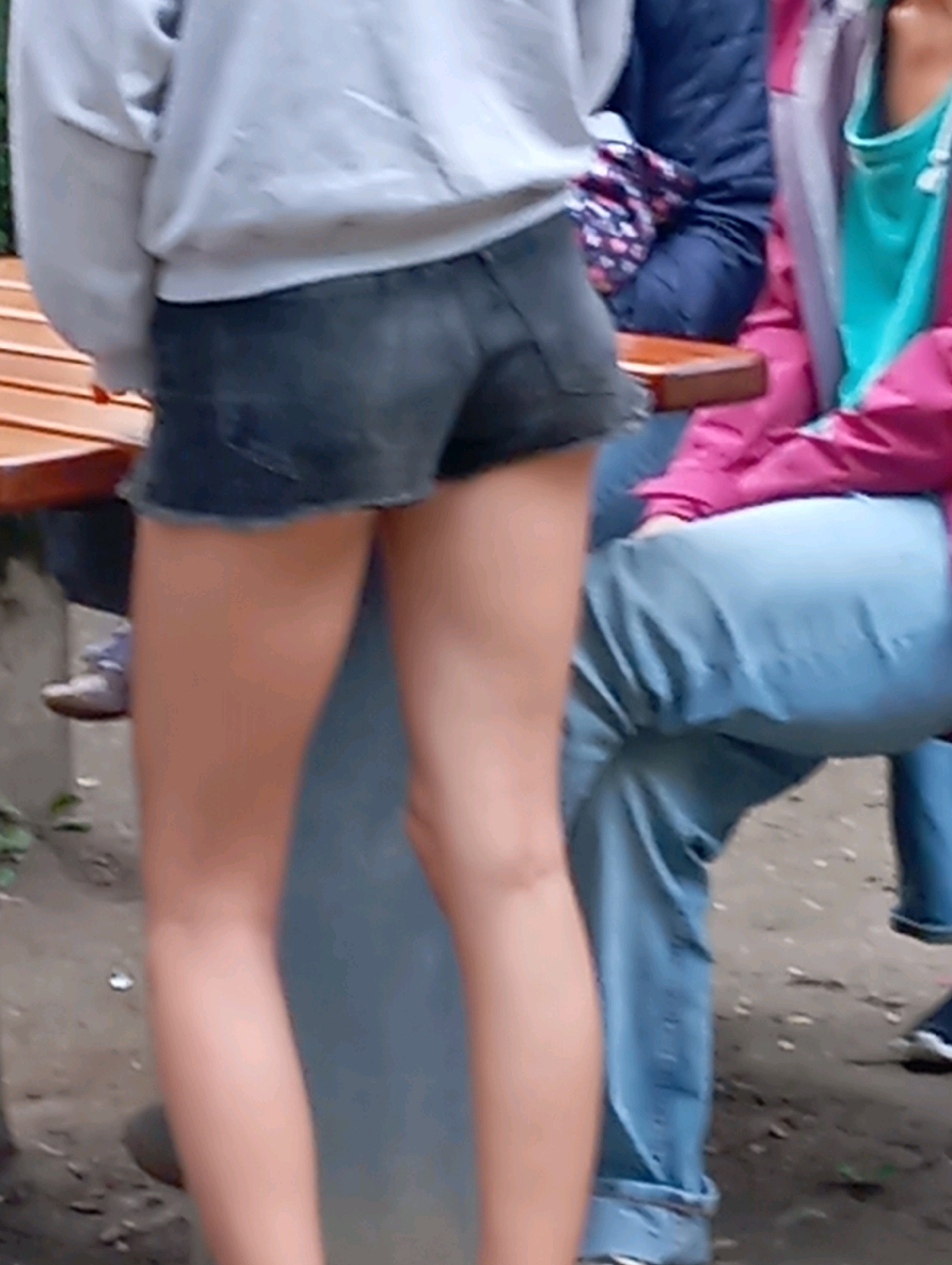 The image shows a close-up of the lower bodies of several individuals outdoors, likely in the city of Baden, Austria. In the foreground, a person wearing dark denim shorts and a light-colored sweatshirt is visible from the waist down. To the right, another individual is wearing light blue jeans and a bright pink jacket over a turquoise top. A third person, partially obscured, is wearing dark blue pants and a patterned item of clothing is visible over their lap. They are seated, suggesting a public seating area, possibly a park or outdoor cafe. The ground appears to be a mixture of dirt and possibly gravel. The overall scene suggests a casual outdoor gathering.
