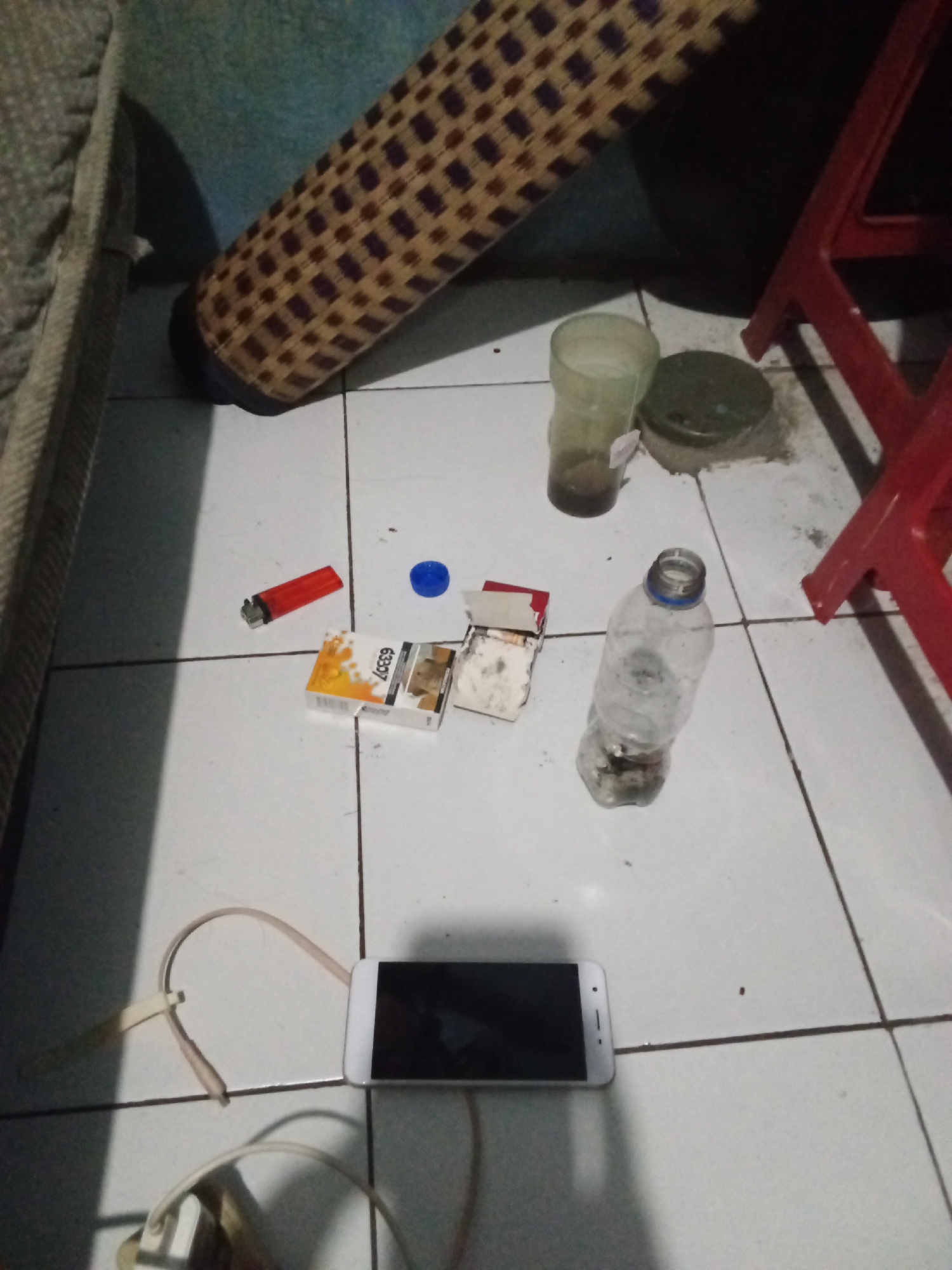 The image depicts a tiled floor in what appears to be a residential setting, possibly a dorm room or a modest living space, in Depok, Indonesia. The scene is cluttered with various items commonly associated with smoking and personal use.
Dominating the foreground is a white smartphone, lying face up, connected to a power source via a white cable. The screen is dark, indicating it's either off or in a sleep state. To the left and below the phone, the end of a beige colored headphone cable is visible, with a white earpiece attached.
Scattered on the floor are remnants of smoking materials. A red lighter lies next to a partially opened pack of cigarettes, labeled "63397". One cigarette appears to have been removed, and several loose cigarettes and what looks like ash are visible within the pack and on the floor. A small blue cap is also present, possibly from a bottle.
Further back, a clear plastic bottle, containing some dark liquid or debris at the bottom, stands upright. To the left of the bottle is a translucent plastic cup containing a dark, murky liquid. Next to the cup is a small, dark, circular object, possibly a lid or container.
In the background, a portion of a rolled-up woven mat is visible, resting against a textured, possibly upholstered surface, likely furniture. A portion of a red plastic chair is also visible on the right side of the image. The lighting suggests it is either evening or the room is dimly lit. The overall impression is one of a personal space with items associated with casual use and perhaps a degree of disarray.