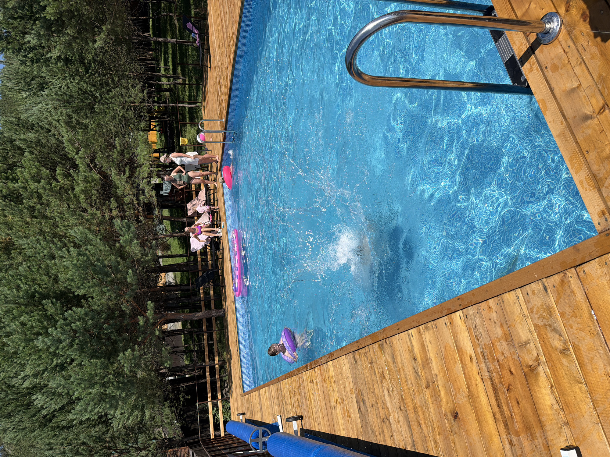 A group of people are enjoying a sunny day at an outdoor swimming pool in Elekmonar, Russia. The pool has bright blue water with ripples and a ladder with chrome rails. A wooden deck surrounds the pool, and there are lounge chairs set up on the deck. In the water, a child with a purple float is splashing, and another person is partially submerged, creating a large splash. Several inflatable toys are floating in the pool, including a pink one. In the background, tall pine trees form a dense forest. Two women and a child are visible on the deck, and two more people are seated on lounge chairs. The overall scene depicts a relaxed, recreational atmosphere.