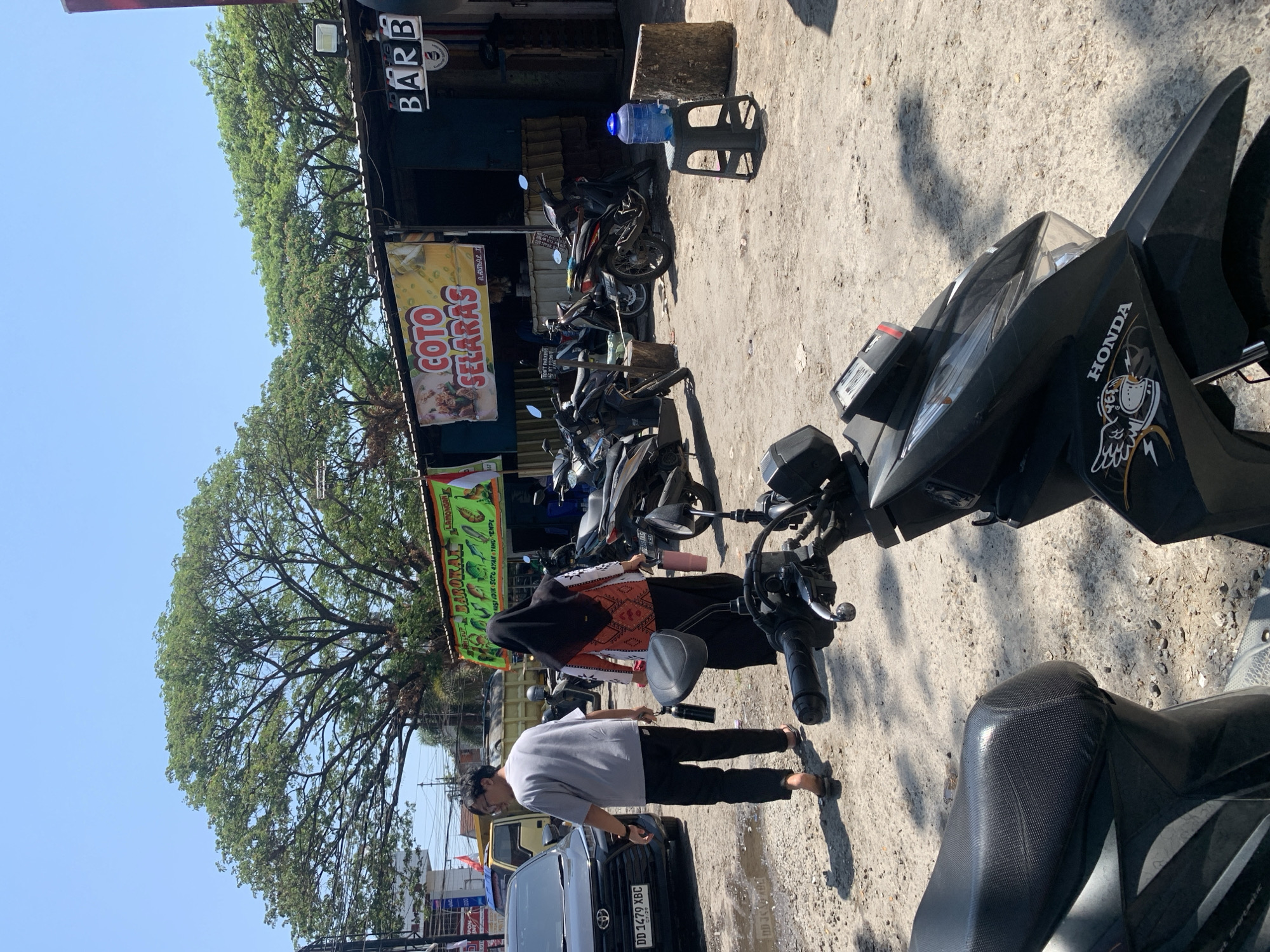The image depicts an outdoor scene, likely a marketplace or street food stall area, in Makassar, Indonesia. The foreground features a black motorcycle, with its front portion clearly visible. Behind the motorcycle, a dirt and gravel ground is visible, with shadows cast from the overhead structure and trees.
In the middle ground, a man in a gray t-shirt and black pants is walking, carrying a black bottle in his left hand. He is barefoot. A woman in a black hijab and a patterned orange and white top is walking ahead of him, carrying a pink bottle. Several motorcycles are parked behind them, along with wooden stumps.
A covered food stall is visible in the background, with banners displaying text, one in yellow with "COTO SELARAS" and another in green with what appears to be a menu. A sign with "BARB" is also visible. A large tree with dense green foliage dominates the upper part of the image, with a clear blue sky above. A yellow truck is parked to the left, next to a dark-colored car with a license plate reading "DD 1479 XBC". A blue water dispenser jug is on a black stool to the right. The overall impression is a busy, casual urban environment.