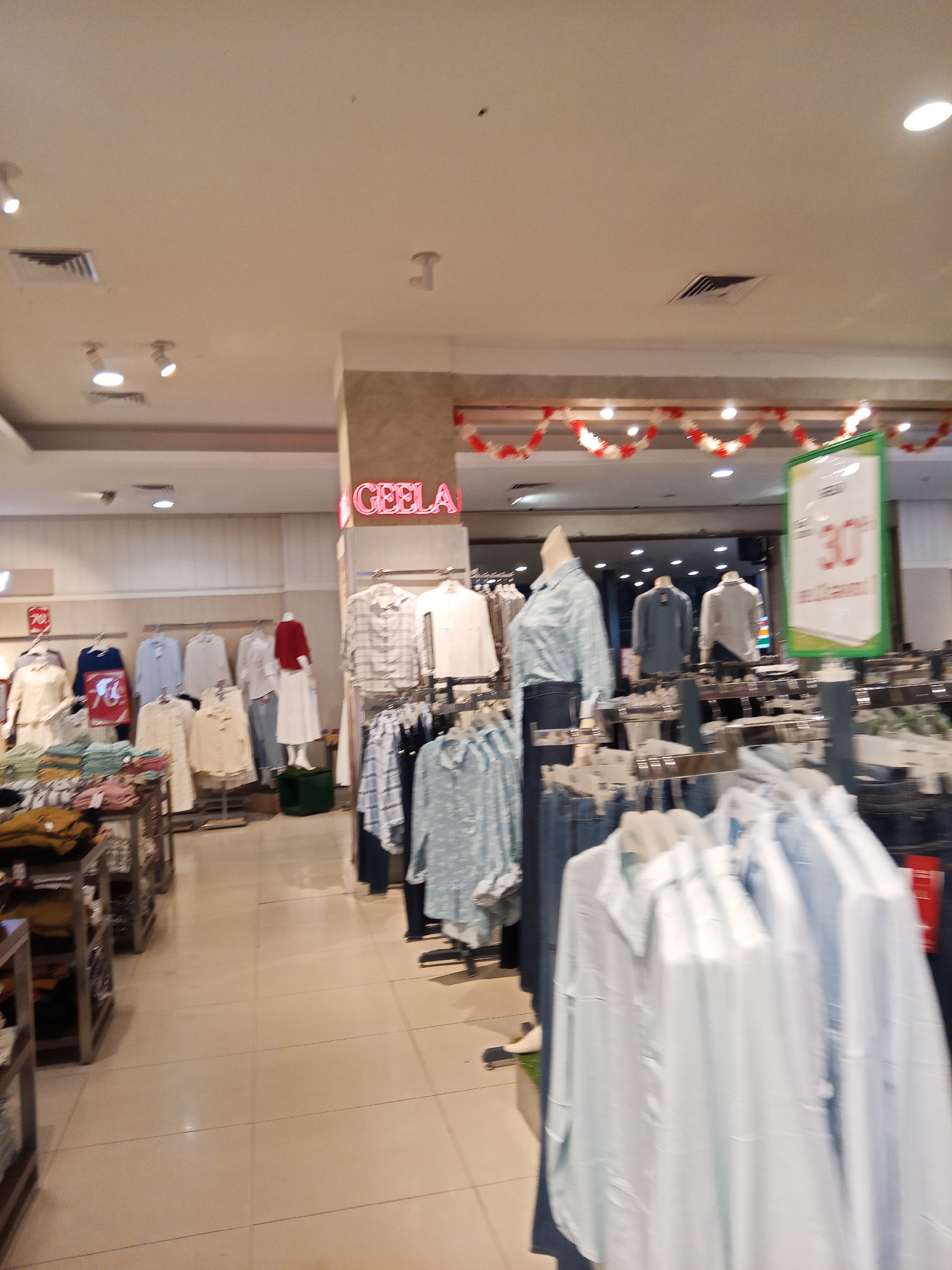 The image depicts the interior of a clothing store, likely a department store, in Lubuklinggau, Indonesia. The store features racks of clothing displayed on mannequins and hangers, with a variety of shirts, blouses, and pants visible. The store has a bright, modern aesthetic, with white ceilings, tiled floors, and fluorescent lighting. A prominent sign with the word "GEELA" in neon pink is visible, indicating the store's name or a brand. Another sign displays "30%" suggesting a sale or promotion. Red and white garlands are strung across the ceiling, possibly indicating a festive occasion or seasonal decoration. The overall scene is a typical retail environment designed to attract customers and showcase merchandise.