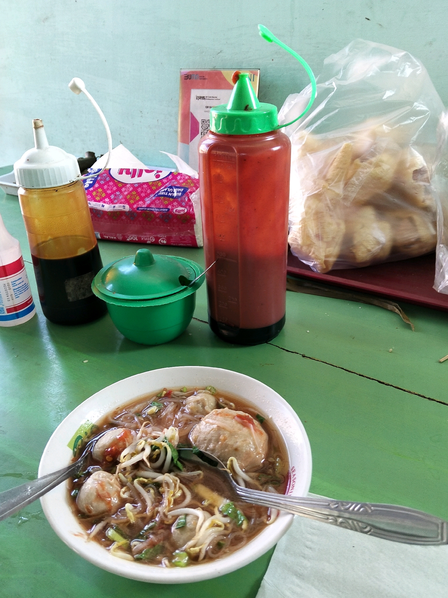 A bowl of Indonesian meatballs (bakso) sits on a green table, accompanied by various condiments and a bag of snacks. The bowl, white and oval-shaped, contains meatballs in a savory broth, garnished with bean sprouts and green onions. Two spoons rest on the rim of the bowl. To the left, a glass bottle with a yellow liquid and a white cap is visible. Next to it, a green condiment bowl with a lid holds a spoon. A tall, red squeeze bottle with a green cap, likely chili sauce, stands to the right of the bowl. Behind these items, a package of pink tissues with a floral pattern is partially visible. A clear plastic bag filled with fried snacks, possibly kerupuk, rests on a dark red tray in the background. The scene is set against a light blue wall. This image depicts a common street food setting, typical of Indramayu, Indonesia, where such meals are frequently enjoyed. The lighting suggests it is daytime, possibly outdoors or in a well-lit eatery. No people are present in the frame.