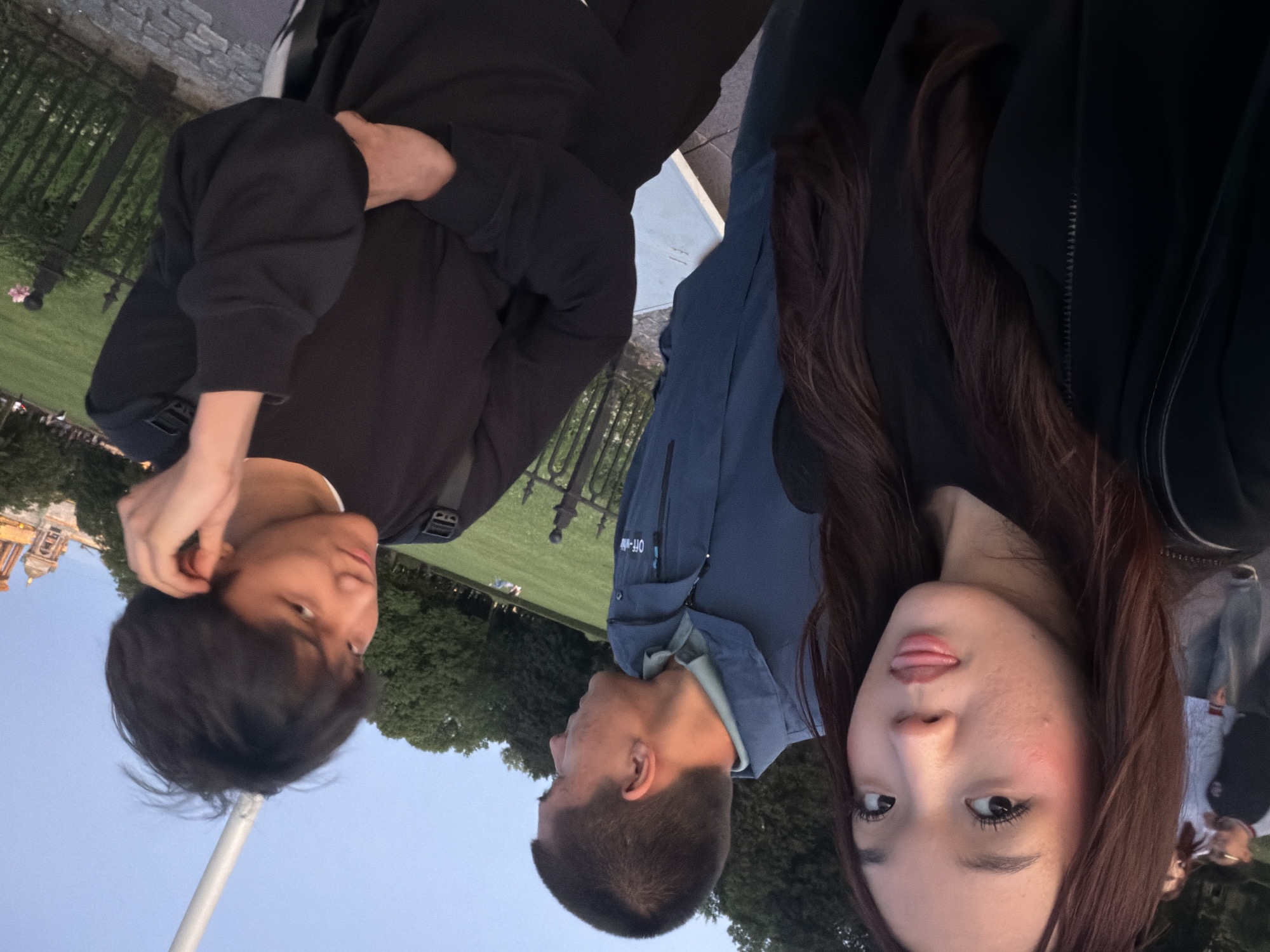 Three people are visible in this vertically oriented, slightly tilted selfie taken outdoors. Two individuals are closer to the camera, with one woman's face prominently featured in the foreground, looking directly at the camera with her tongue slightly out. She has dark hair and is wearing a dark jacket. To her left, a man is also facing the camera, with his hand near his ear. He has short dark hair and is wearing a black sweatshirt. Behind them, a third person, a man wearing a blue jacket with "OFF-WHITE" and other text visible on it, is partially obscured.
The background shows a grassy area with trees and a black metal fence. A building with a domed top and classical architecture is visible in the far left background, suggesting a potentially historic or significant location. The sky is a clear, pale blue, indicating daytime. The lighting suggests it might be late afternoon or early evening. The overall mood appears casual and lighthearted, given the playful expression of the woman and the casual pose of the man. The presence of the building and the general scenery are consistent with a location in Saint Petersburg, Russia.