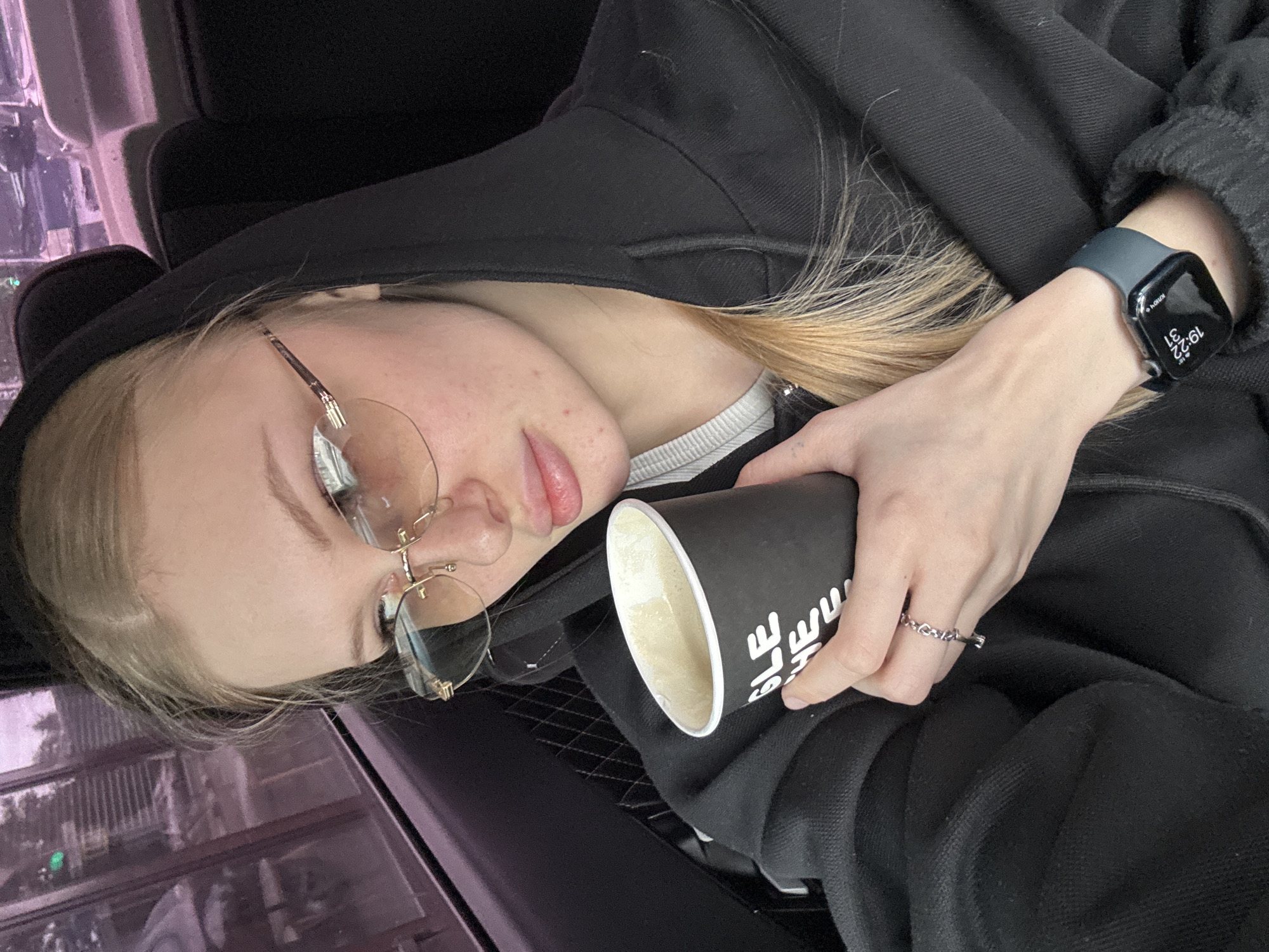 A young, light-skinned person with blonde hair and round glasses, wearing a black hooded sweatshirt, is seated in what appears to be a vehicle. They are holding a black cup with white lettering. The person is wearing a dark-colored smartwatch on their left wrist and a silver ring on their left hand. The timestamp on the watch is 19:22. The background shows reflections of buildings through the window, suggesting an urban environment. Considering the prompt's mention of Perm, Russia, it can be deduced that the scene is likely within that city.