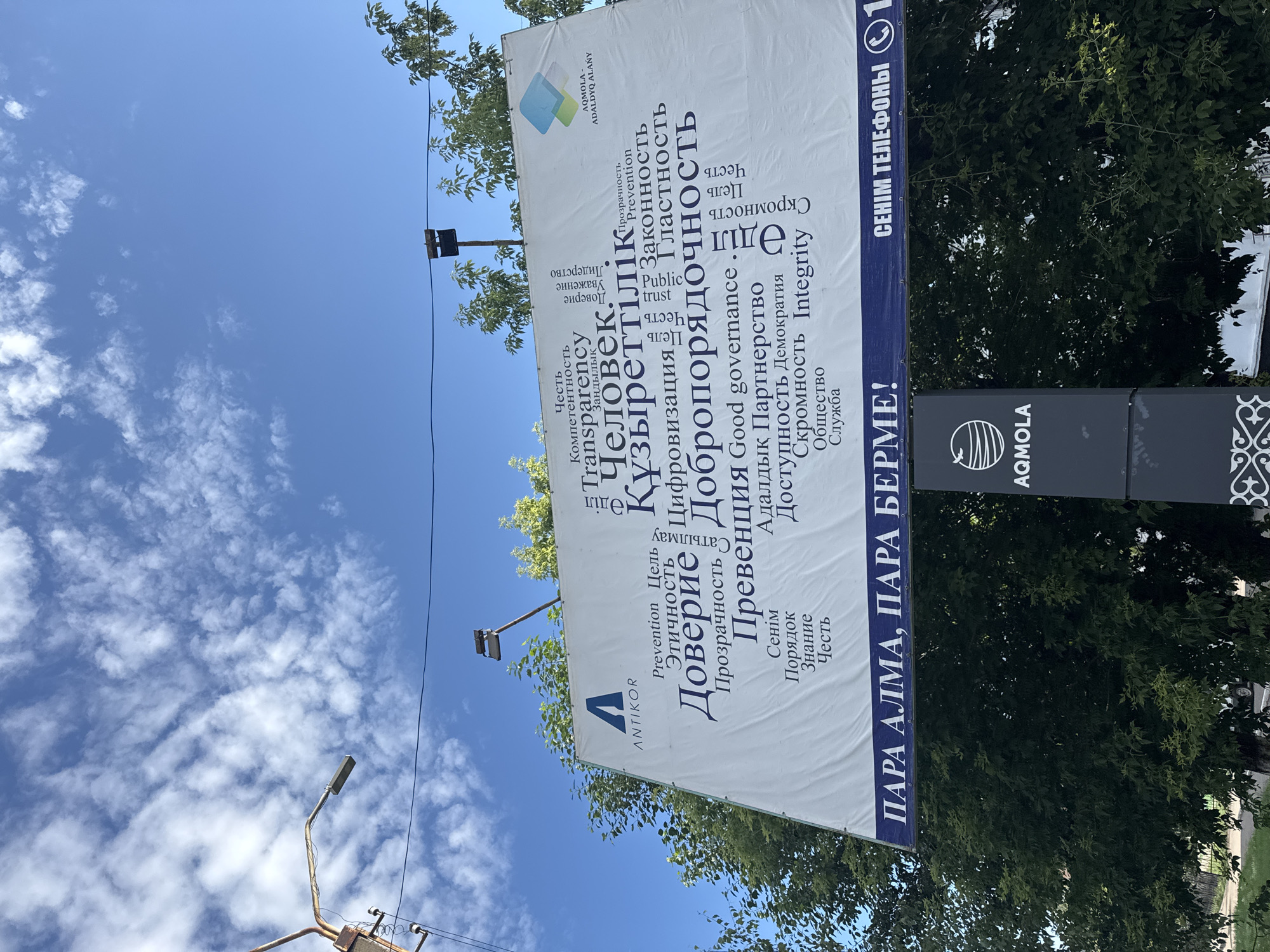 The image shows a large outdoor billboard in Kokshetau, Kazakhstan, displaying a word cloud with various terms in Kazakh and Russian, along with the logo of "ANTIKOR" and "AQMOLA-ADALDVQ ALANY". The billboard is mounted on a white surface and is partially obscured by trees. Below the billboard, there is a dark blue banner with white text that reads "PARA ALMA, PARA BERME!" on the left and "SENIM TELEFONY" followed by a phone icon and the number "1" on the right. To the right of the billboard, a dark vertical structure with the "AQMOLA" logo is visible. The sky is blue with scattered white clouds. A street lamp is visible in the upper left corner of the image. The scene appears to be an outdoor public space.