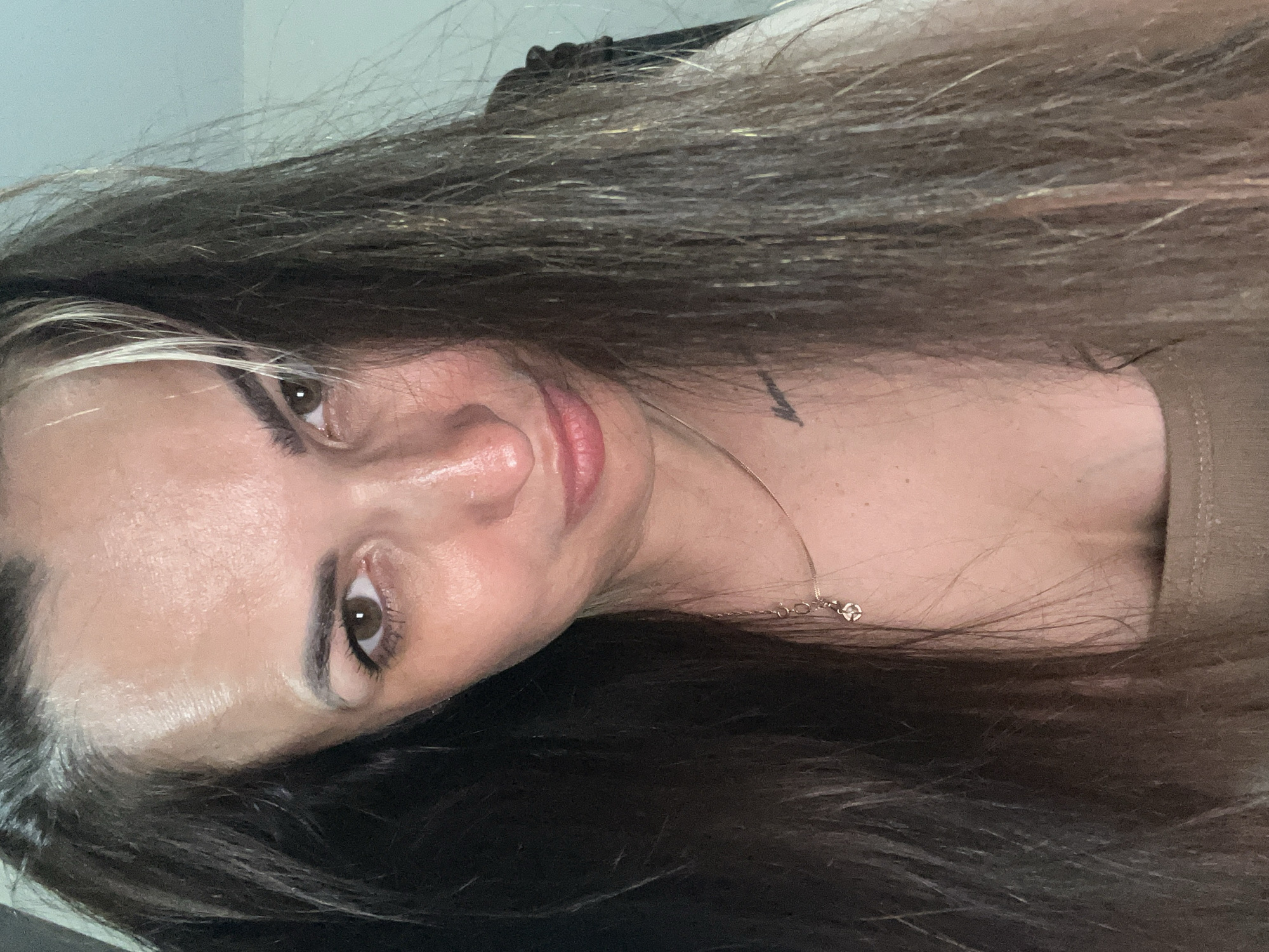 The media file is a close-up, rotated photograph of a woman's face and upper chest. She has long, dark brown hair with lighter streaks, which frames her face. Her eyes are a warm brown, and she is looking directly at the camera with a neutral expression. Her lips are slightly parted and appear to be colored with a pink lipstick. She is wearing a thin necklace with a small, delicate pendant. A hint of her shoulder and upper chest are visible, where she is wearing a brown garment. There is a faint tattoo visible on her neck. The lighting suggests an indoor setting, and the time of day is not discernible. The overall mood is intimate due to the close-up framing.