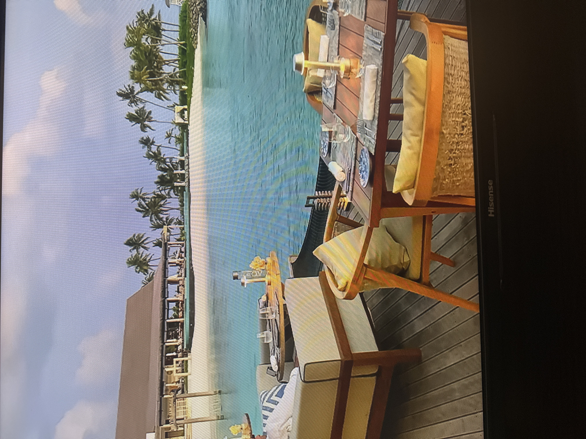 This image shows an outdoor dining area at a tropical resort. There are several tables and chairs set up on a wooden deck overlooking a body of turquoise water. In the background, there is a building with a thatched roof, palm trees, and a sandy beach. The water appears calm and clear. The lighting suggests it is daytime, possibly late afternoon. There are no people clearly visible in the frame. The scene depicts a serene and luxurious vacation setting. The image is not located in the city of Kyiv, Ukraine.