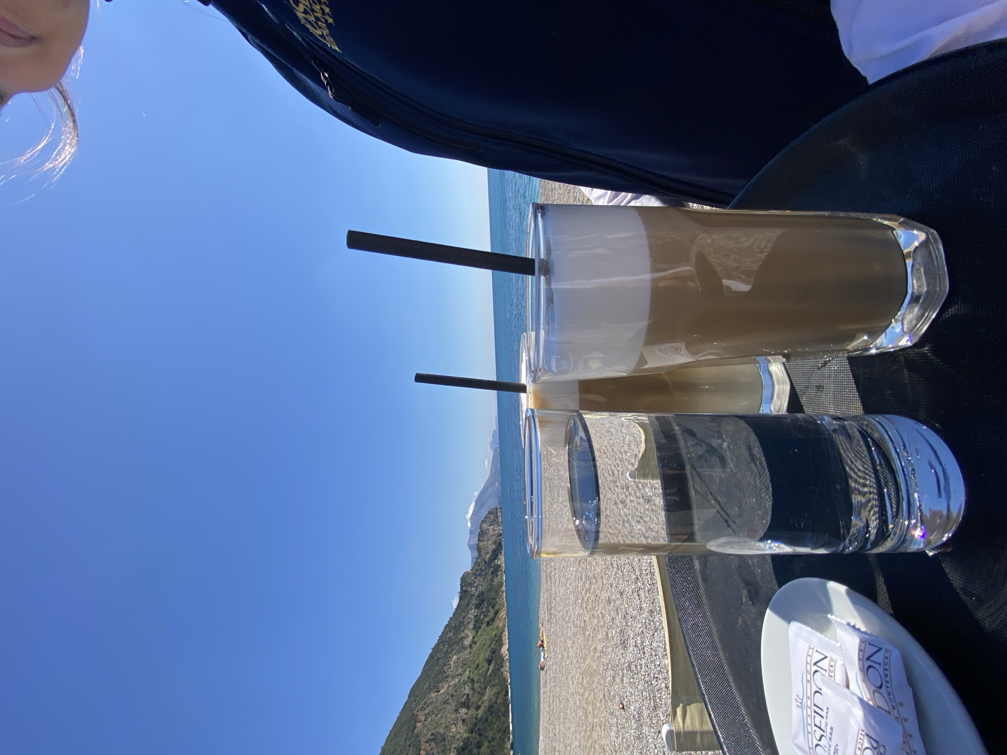 The image captures a scene at a beachfront location, likely in or near Prijevor, Montenegro, based on the hotel branding on the small white plate that reads "Beach Hotel Poseidon Rose / Montenegro." The foreground features two tall glasses with black straws; one appears to contain a layered coffee drink with a foamy top, while the other holds clear water. They are positioned on a black surface with a gray grid pattern, possibly a table or chair. The background showcases a picturesque coastal landscape. The Adriatic Sea stretches to the horizon beneath a clear blue sky. To the left, a steep, green hillside descends towards the beach, suggesting a mountainous coastal region. The beach itself is visible and looks like it consists of pebbles and gravel. The edge of a person’s face and hair is visible at the top left corner of the frame. In addition, the top of a dark bag or other personal item can be seen at the top of the image, which might be located on a table next to the beverages.