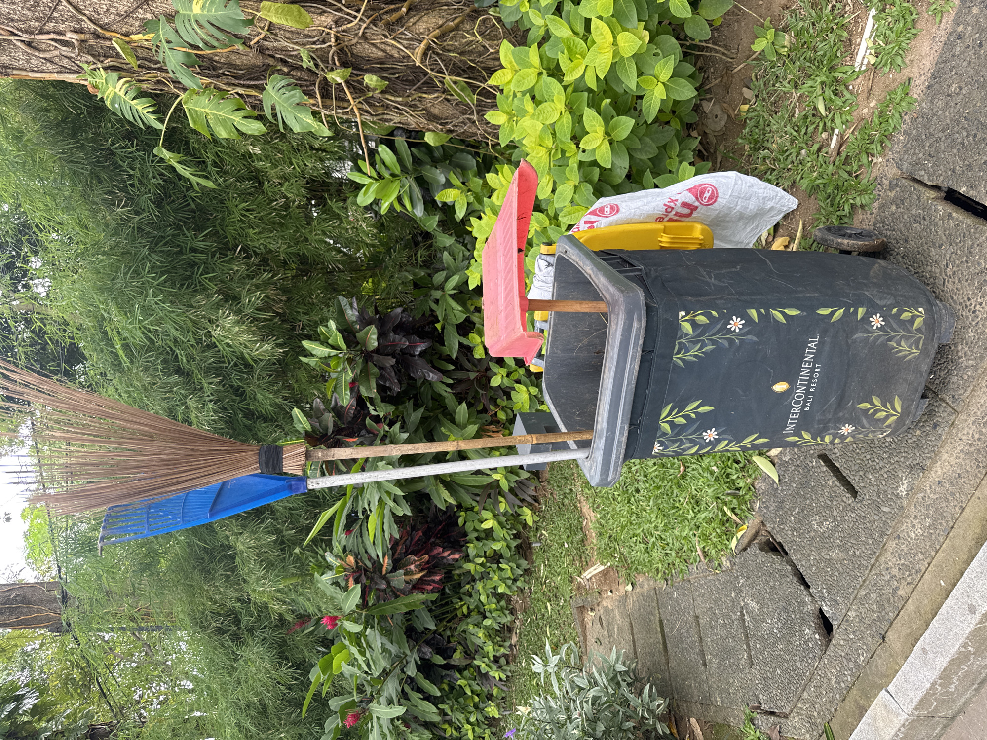 A black rolling trash can with the text "InterContinental Bali Resort" printed on its side sits on a stone-paved surface next to lush green foliage. A broom and a dustpan are leaning against the trash can. A red shovel and a yellow container are partially visible inside the can, with a white shopping bag next to it. The location is Kuta Selatan, Indonesia, as indicated by the trash can's branding.