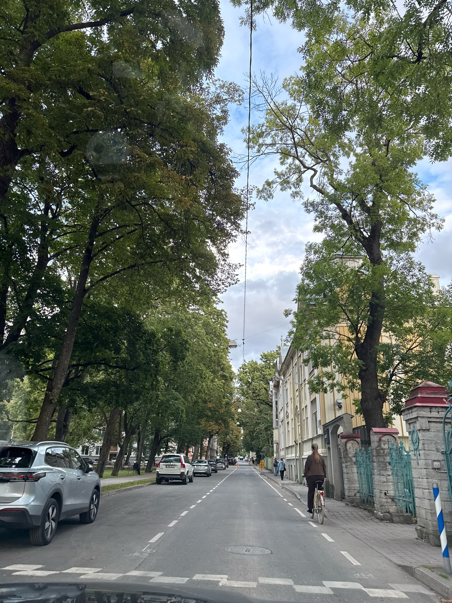 The image shows a street scene in Tallinn, Estonia. In the foreground, a silver SUV is parked on the left side of the road. The street is paved with asphalt and has white lane markings. A pedestrian crossing is visible in the foreground. Further down the road, there is a person riding a bicycle in the right lane. On the right side of the street, there are buildings with light-colored facades and ornate architectural details, as well as a stone wall with a green decorative fence. Both sides of the street are lined with mature trees that cast shadows on the road. The sky is partly cloudy. The overall impression is of a quiet, tree-lined urban street on a cloudy day.