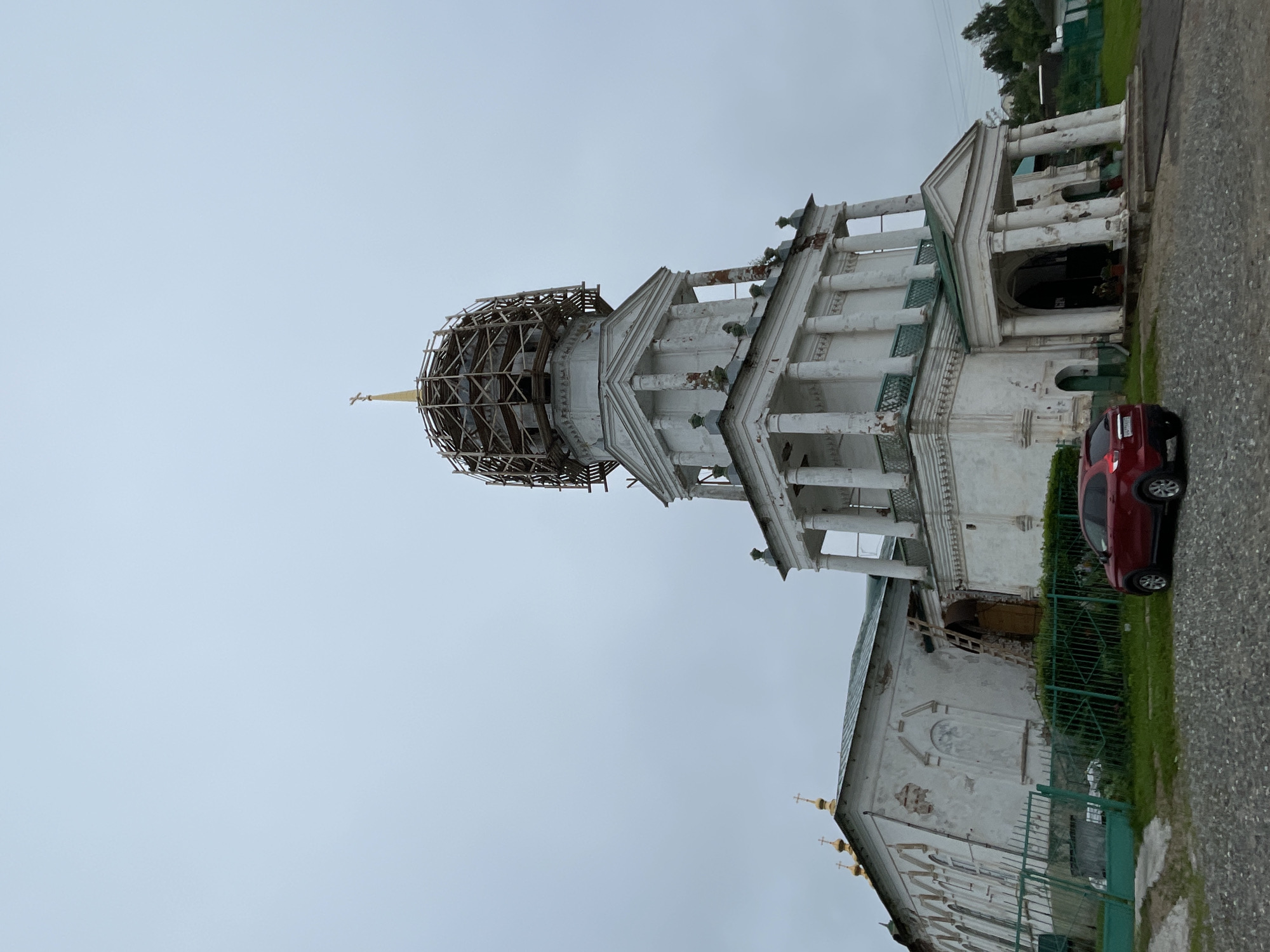 The image shows a white, multi-story church with a green roof and a golden cross on top. The church appears to be under renovation, as scaffolding is visible around the upper dome. A red car is parked in front of the church. The sky is overcast. The church is located in the village of Кстинино, Russia.