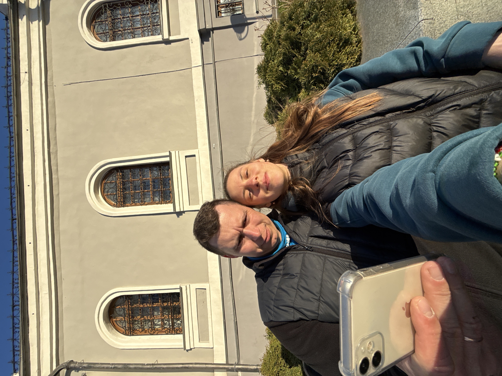 A man and woman are taking a selfie in front of a light grey building with three arched windows containing decorative ironwork. The building has white architectural trim. The man is wearing a black puffer jacket over a blue shirt. The woman has long brown hair and is also wearing a black puffer jacket. The man is holding a smartphone to take the picture. A green bush is visible next to the building. The location is Kazan, Russia.