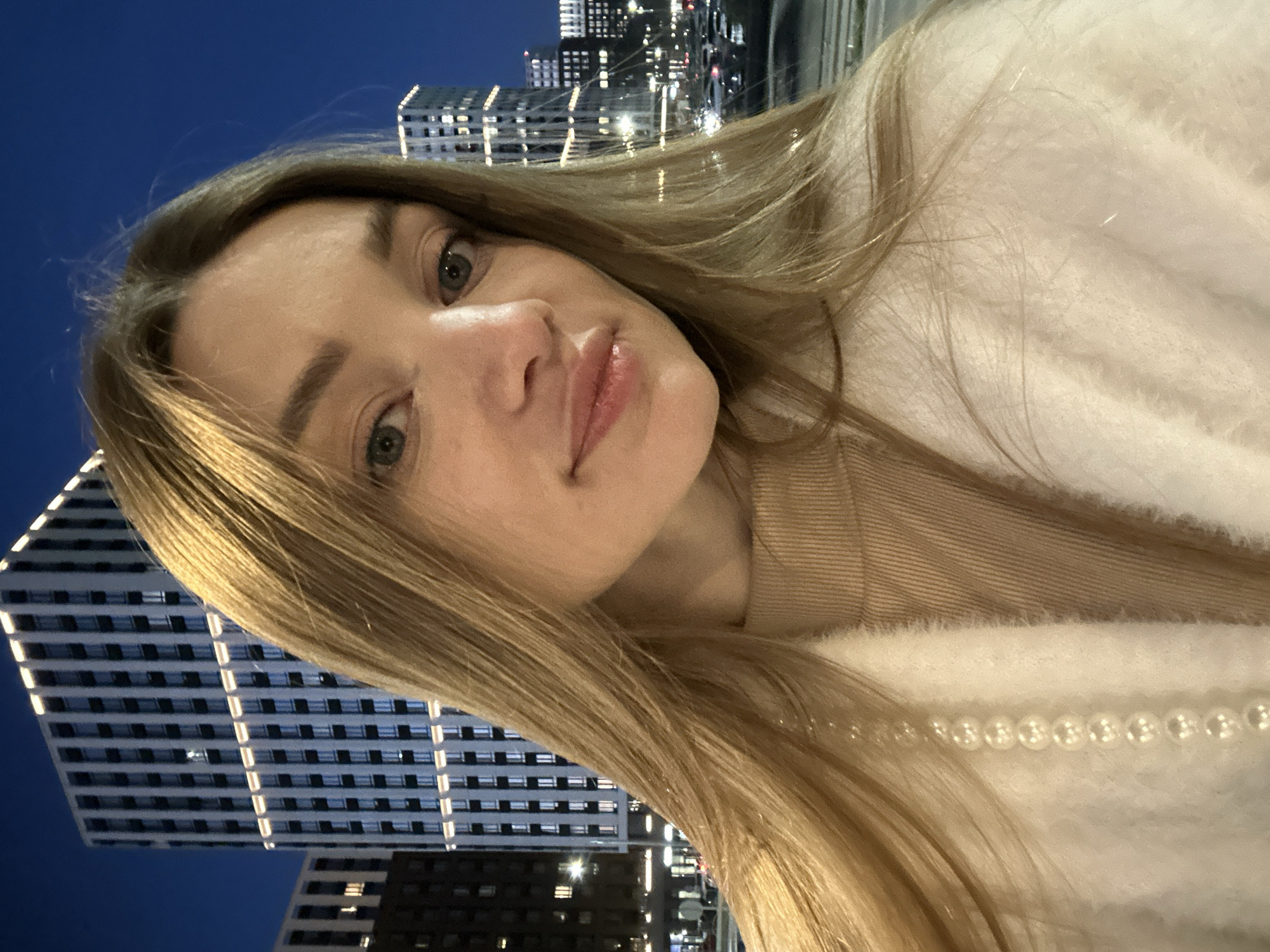 A medium close-up, rotated selfie captures a young woman looking directly at the camera with a calm expression and a subtle smile. She has long, straight light brown or blonde hair, and blue-grey eyes. She is wearing a light, possibly cream-colored, fluffy sweater or jacket over a beige top. A string of pearls is visible draped over her right shoulder and the light-colored garment.
The background reveals a vibrant city skyline at night, featuring multiple modern high-rise buildings with many illuminated windows arranged in grid patterns. The sky is a deep blue, indicating either late dusk or nighttime. The perspective suggests the photo was taken from a lower angle, looking upwards towards the buildings. The setting is an urban environment, identified as Kazan, Russia by the provided context. No visible text is present in the image.