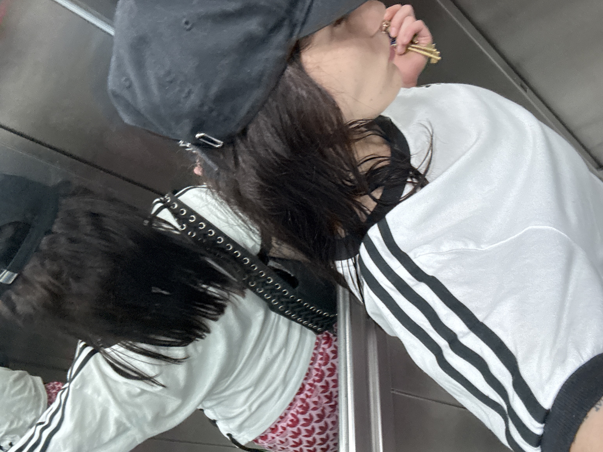 A person wearing a black cap and a white shirt with black stripes is visible in a slightly tilted perspective, likely a selfie taken in a mirror. The person's hair is dark, and they are wearing rings on their fingers. A black belt with silver eyelets is visible. The mirror reflects the same person wearing a pink and white leaf patterned shirt underneath the white shirt. The background appears to be an elevator interior, with a reflective metal surface.
There is no information present in the image to determine if this was taken in Buenos Aires.