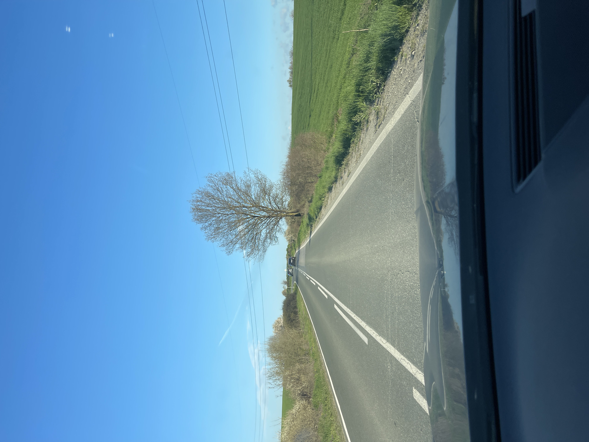 A view from inside a vehicle traveling along an asphalt road in a rural setting. The sky is blue, and there is a tree with bare branches on the left side of the road. Power lines run parallel to the road. A green field borders the road on the right, along with some low bushes. Other vehicles are visible in the distance. The exact location within Burgas, Bulgaria, cannot be determined from the image.