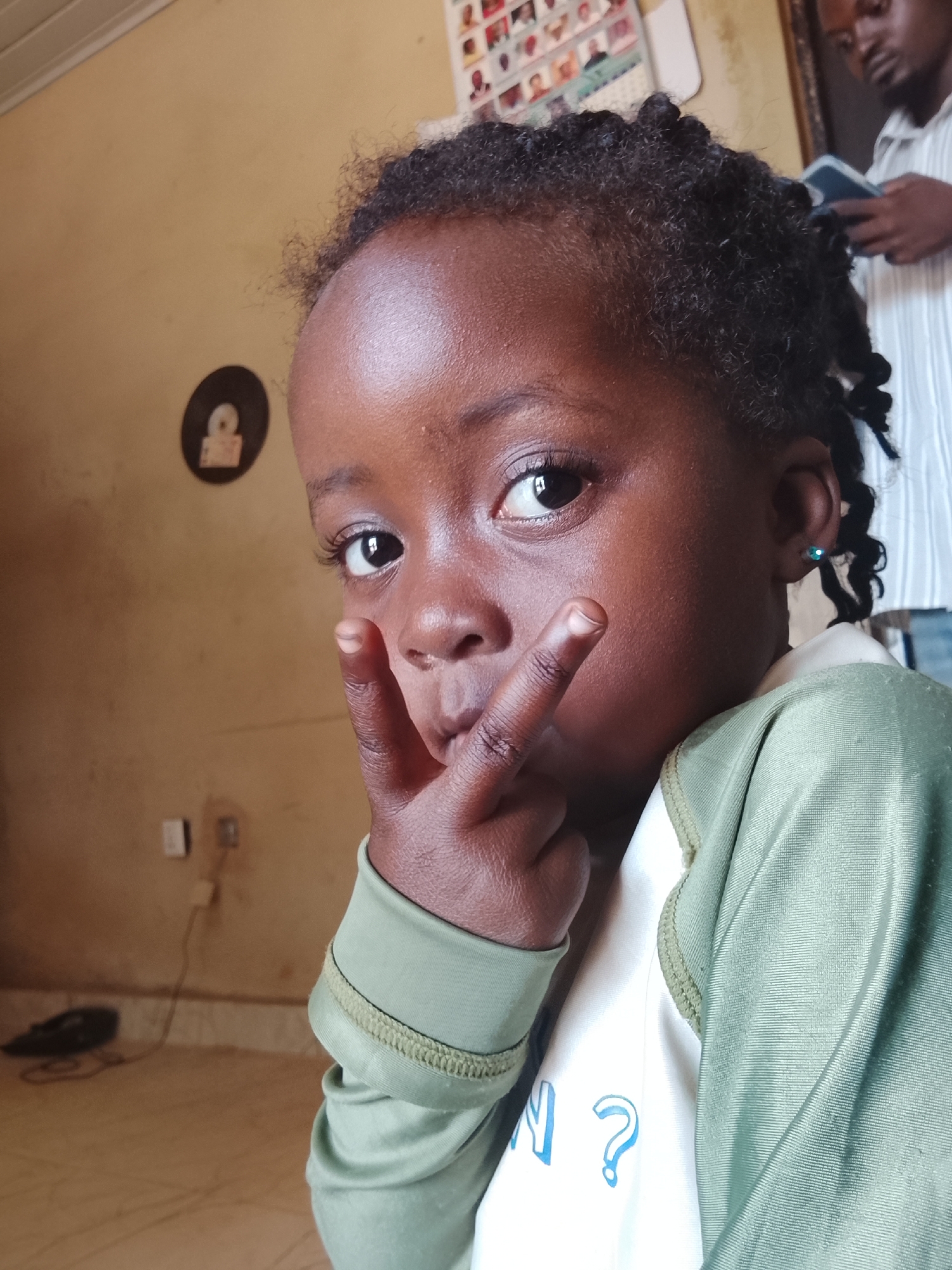 The image features a young, dark-skinned child making a "peace" or "victory" sign with their fingers. The child has dark, curly hair styled in small twists and a blue earring in one ear. They are wearing a green long-sleeved shirt over a white shirt with blue writing partially visible, including a question mark. The background includes an adult male with a dark complexion, partially visible on the right, looking down at a smartphone. The wall behind the child is beige with a circular object attached, possibly a sticker or decoration. An electrical outlet and a shoe are also visible in the lower left corner. The location is indicated as Kuru Dungus, Nigeria.