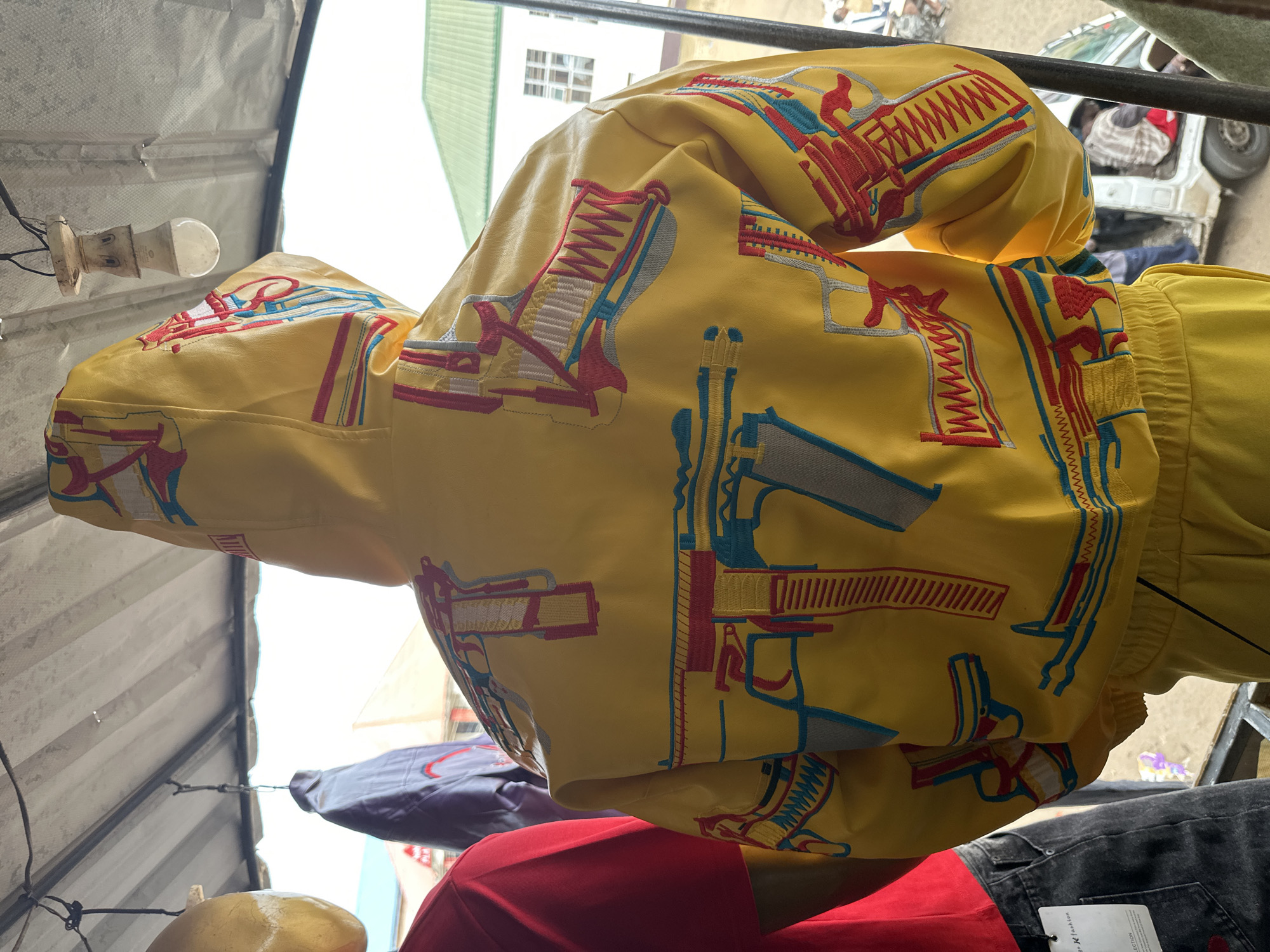 In Bukuru, Nigeria, a yellow hooded jacket with a gun-themed pattern is displayed on a mannequin. The jacket is adorned with embroidered images of guns in red, blue, and white. The jacket is part of a tracksuit, with the lower portion of yellow pants visible. To the left of the mannequin, a red t-shirt is partially visible, and to the far left, a yellow object, possibly another mannequin's head, hangs. Behind the displayed clothing, a grey coat is hung, and further in the background, a white building with a green roof and windows can be seen. A white vehicle is also visible in the background. The scene appears to be an outdoor market stall, indicated by the corrugated metal roofing overhead and hanging light fixtures.