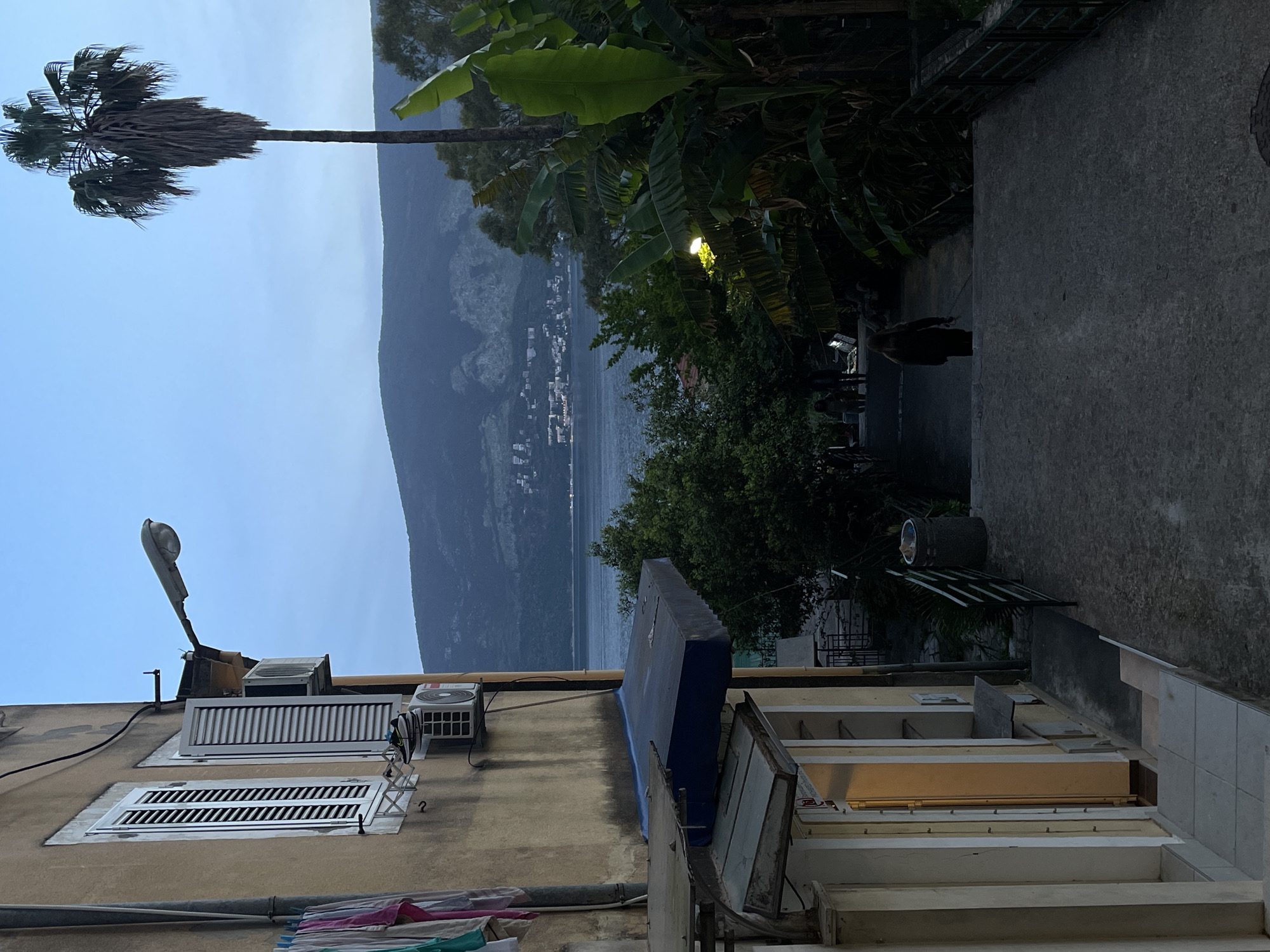 This is an outdoor view, possibly taken from a balcony or elevated position, in Herceg Novi, Montenegro. The scene is oriented toward a landscape with a large mountain or hill across a body of water, likely the Bay of Kotor. The sky is pale blue, possibly at dusk or dawn. A tall palm tree is visible on the left side, extending into the sky. Banana trees and other foliage are growing closer to the viewpoint.
In the foreground, there are buildings with architectural details such as white louvered windows, air conditioning units, and railings. Some laundry is hanging on a line outside one of the buildings. A streetlight is attached to the building in the foreground. The foreground also includes a concrete pathway or terrace. People are moving further in the background near outdoor seating and tables, possibly a restaurant or cafe.