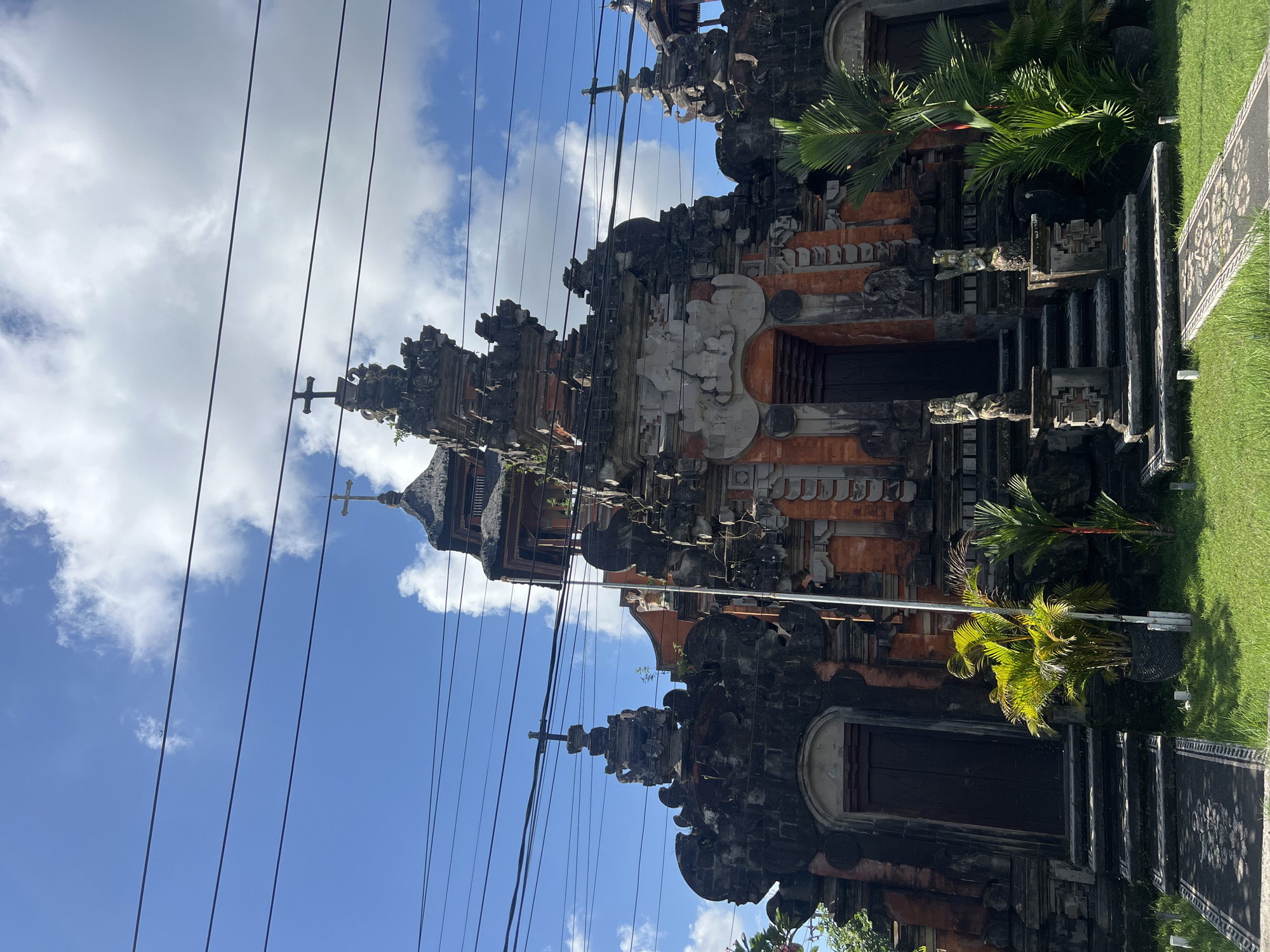 A medium shot captures the ornate facade of a Hindu temple, possibly a church or a temple with Christian influences given the prominent crosses atop its spires, in North Kuta, Indonesia. The temple is constructed from dark stone and reddish-orange brick, adorned with intricate carvings and traditional Balinese architectural elements. Multiple dark wooden doors are visible, some set within arched entrances. A series of steps leads up to the central entrance, flanked by two stone statues of winged figures. Lush green grass covers the foreground, with potted palm trees and other greenery adding to the tropical ambiance. A paved pathway with a decorative mosaic pattern leads towards the temple entrance. Overhead, a network of black electrical wires crisscrosses the bright blue sky, which is dotted with fluffy white clouds. A distant airplane is visible in the sky. The lighting suggests it is daytime, with sunlight illuminating the scene.