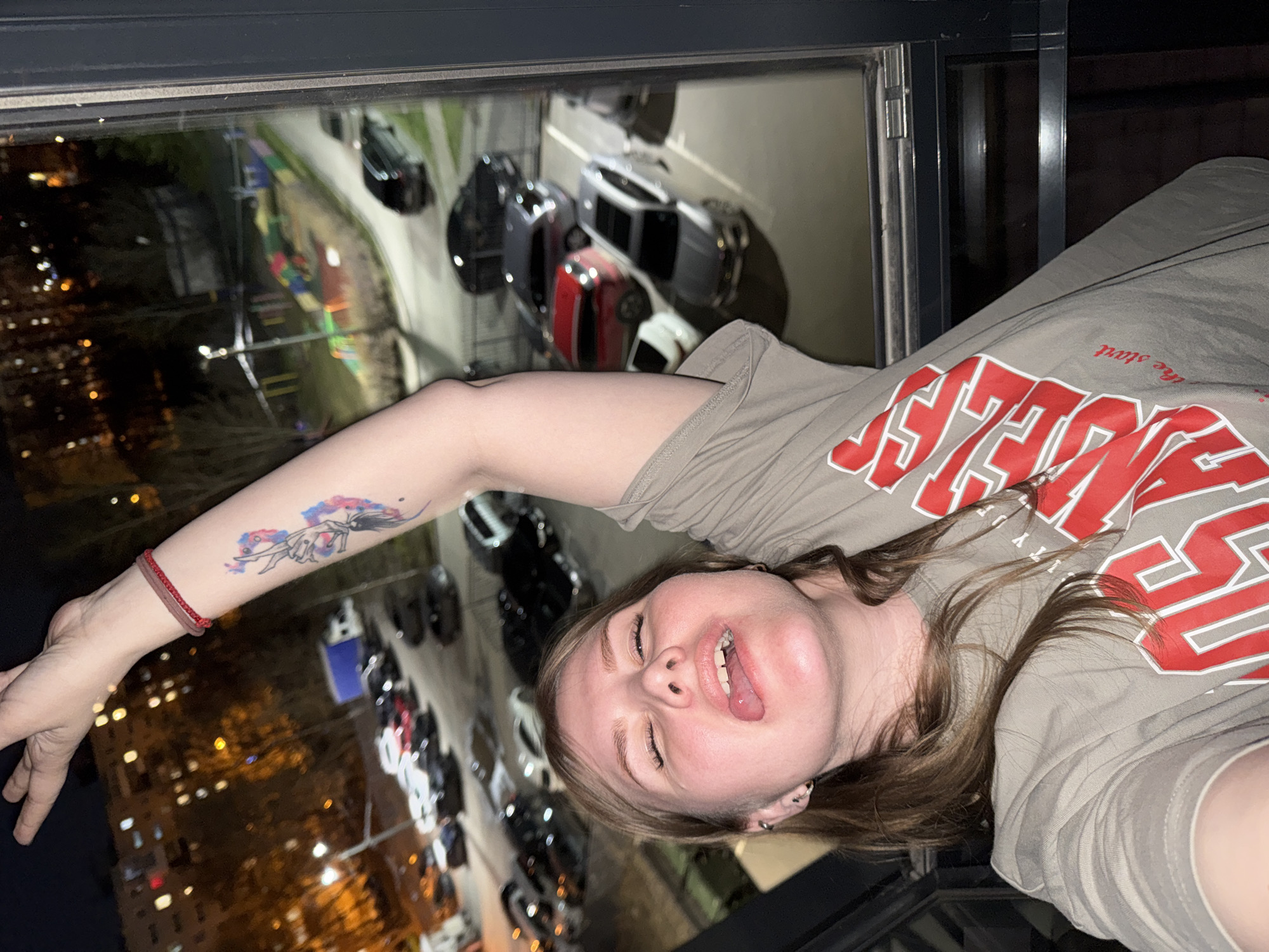 A young woman is captured in a horizontally oriented selfie, seemingly lying down and looking playfully upwards into the camera. Her head is positioned towards the bottom right, with her left arm extended across the frame towards the upper left. She has light brown hair and a light complexion, with her eyes partially closed and her tongue slightly extended in a casual, possibly humorous, expression. She has a nose piercing and multiple ear piercings.
She is wearing a light tan or khaki t-shirt featuring prominent red and white block lettering that reads "GEORGIA," with smaller text below it that says "the start." A colorful tattoo depicting a stylized figure or abstract design is visible on her left forearm, along with a red beaded bracelet on her left wrist.
The background, seen through a large window, is a nighttime cityscape. Out-of-focus bokeh lights from distant buildings and streetlights indicate an urban setting in Perm, Russia. Below the window, several parked cars of various colors (including dark, red, and lighter vehicles) are clearly visible on what appears to be a street or parking lot. The interior frame of the window is visible, suggesting the photo was taken from inside a building overlooking the city.